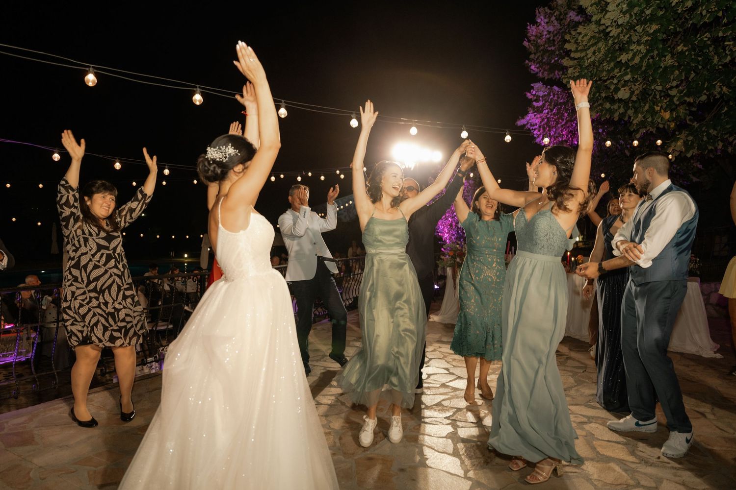 Wedding guests dance and celebrate under twinkling string lights during an outdoor evening reception.