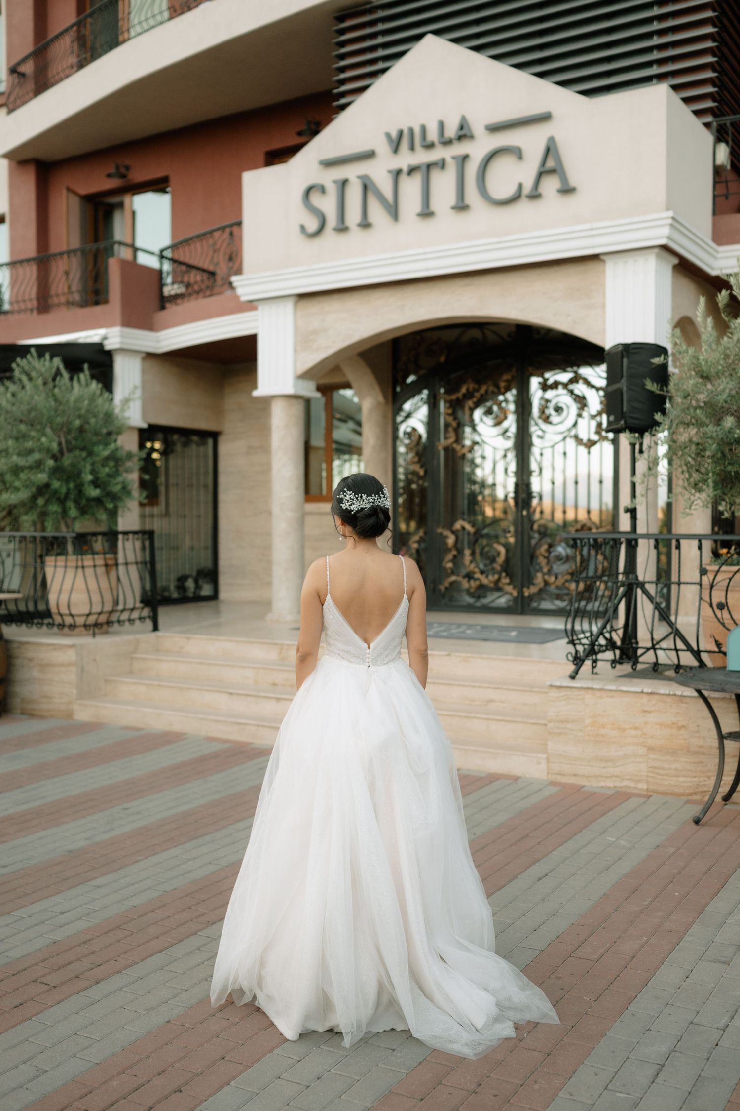 A bride in a flowing white wedding gown stands on a tiled outdoor terrace near a building entrance.