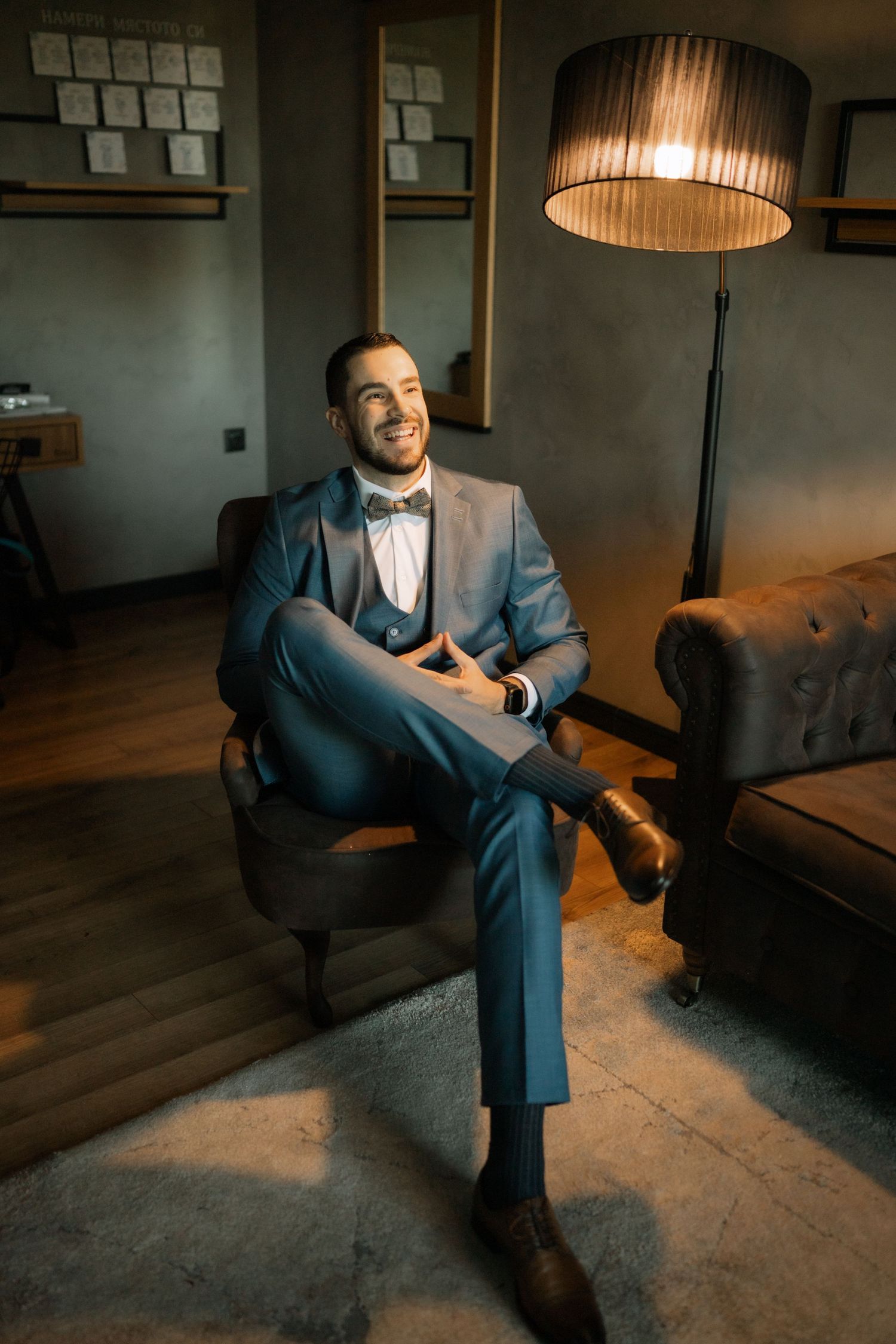 A person in a blue suit sits on a grey couch in a dimly lit room with warm lighting from a pendant lamp above.