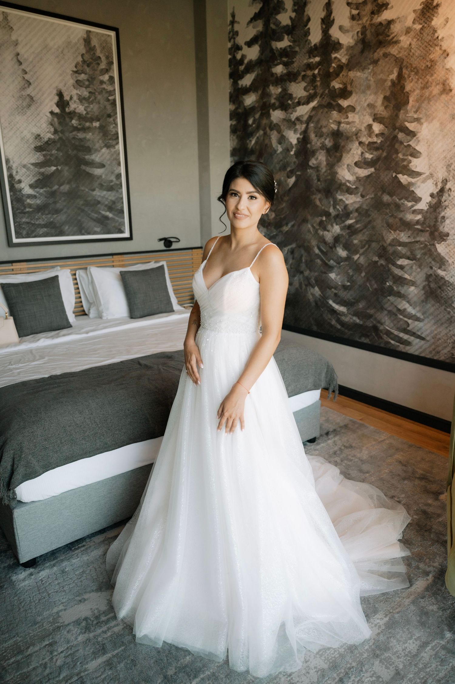 A stunning white wedding gown with spaghetti straps and tulle skirt is showcased in a room with forest-themed wall art.