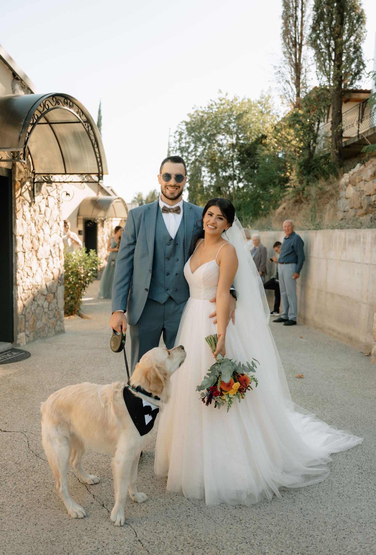 A bride and groom pose with their golden retriever dog on their wedding day in a rustic outdoor setting.