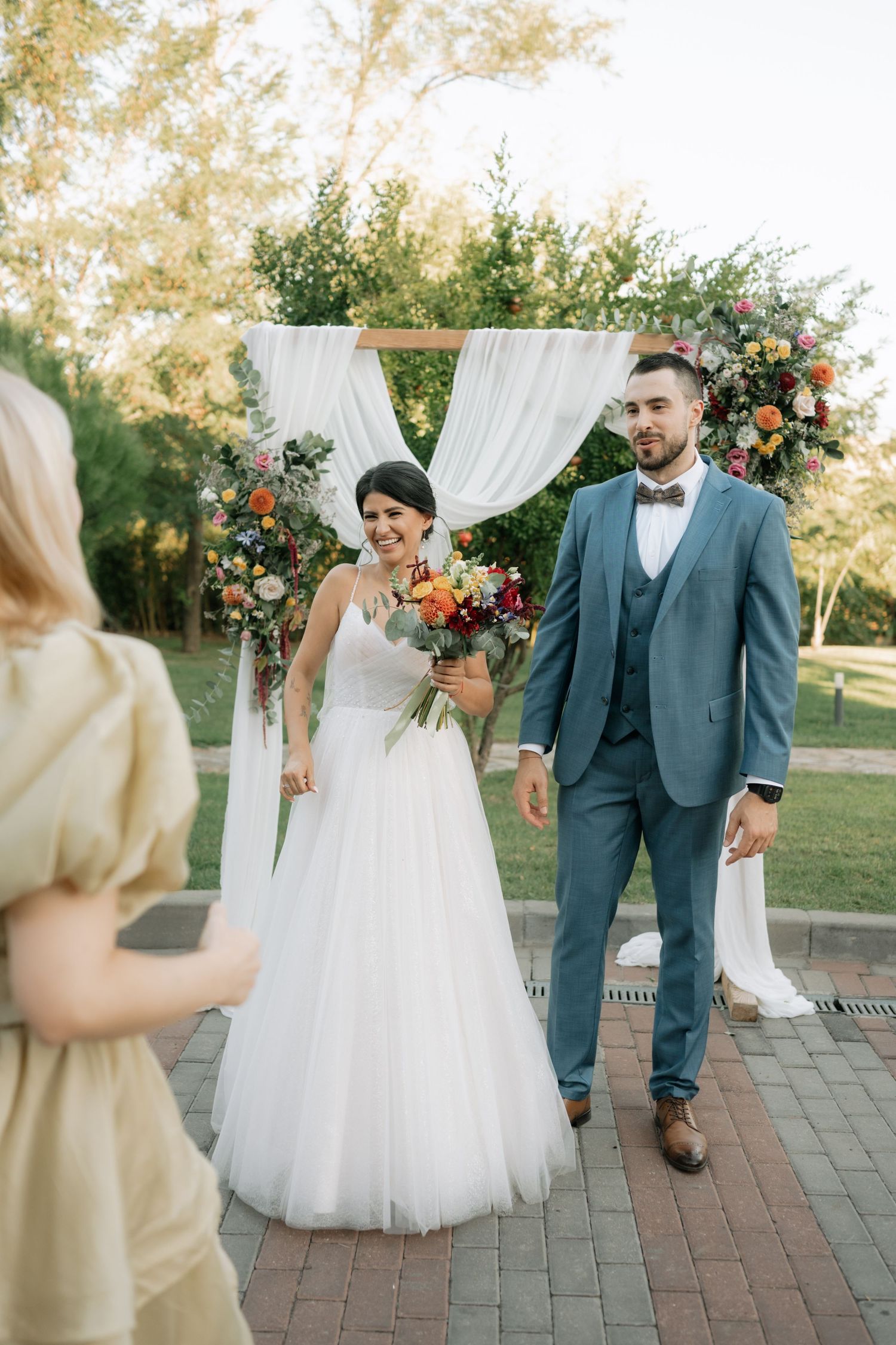 Wedding couple walks together on brick pathway under floral arch decorations at outdoor garden ceremony.