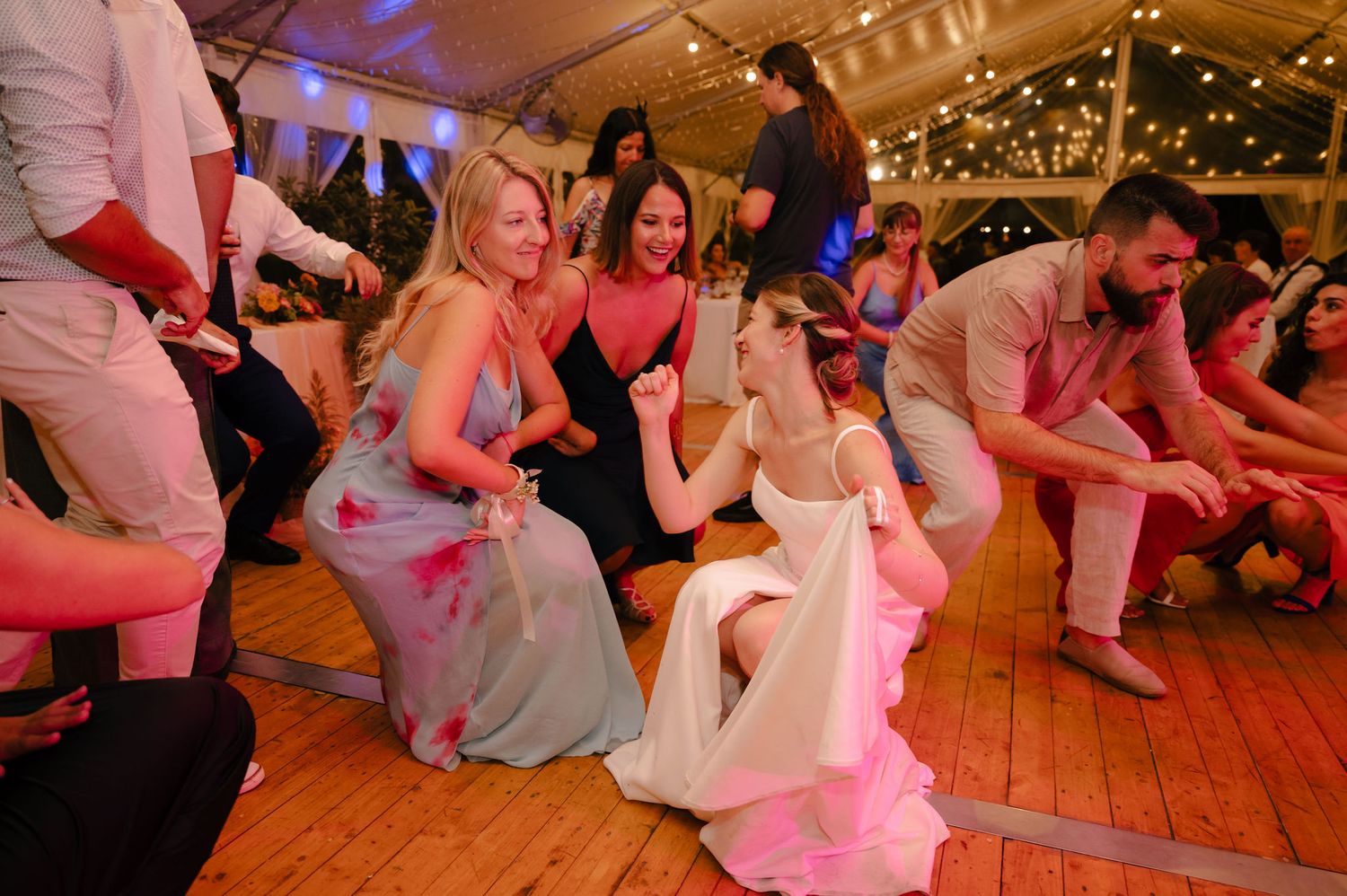 Wedding guests on a wooden dance floor participate in a fun group dance activity under warm lighting and decorations.