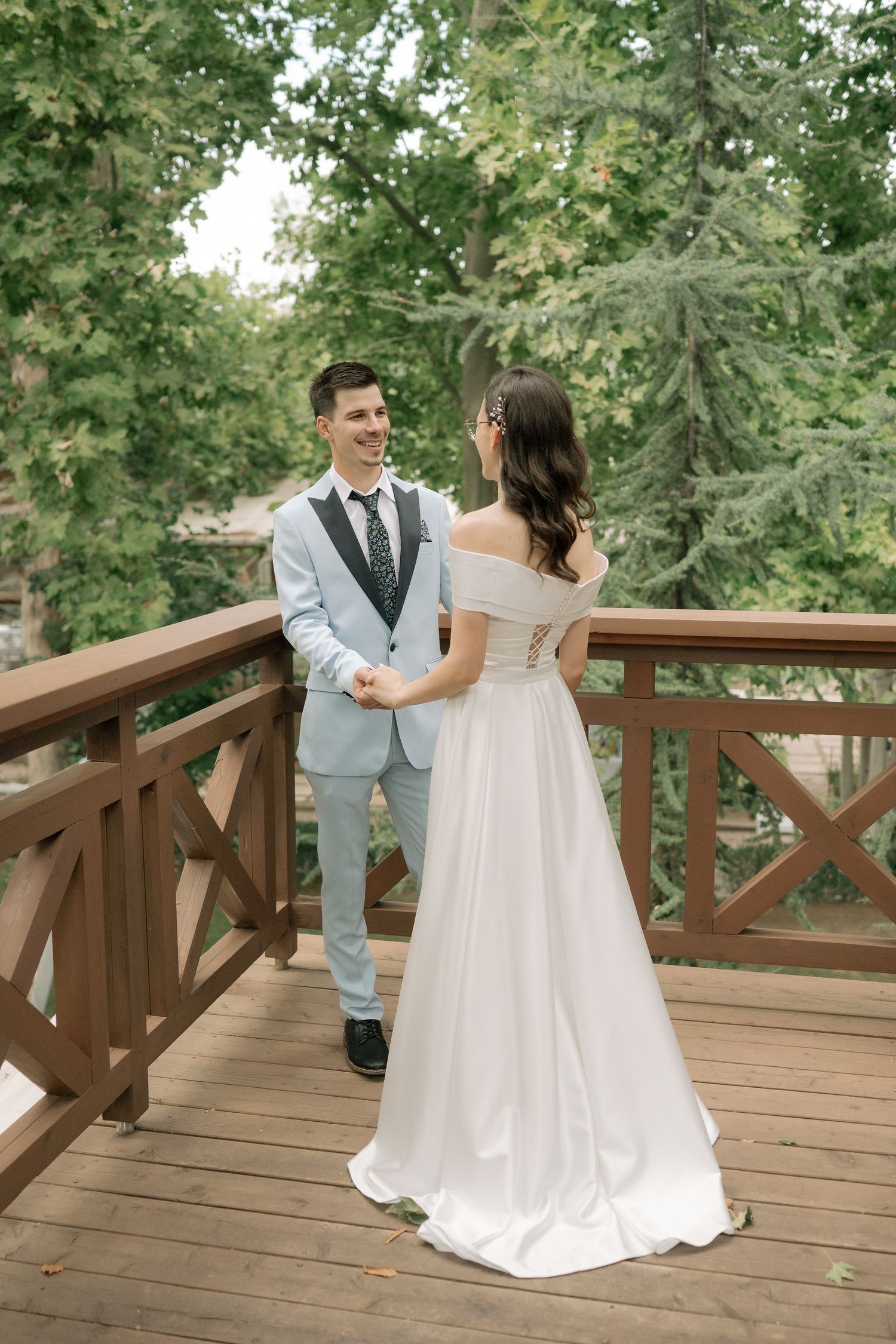 A romantic wedding couple dances together on a wooden deck surrounded by lush green trees in a beautiful outdoor setting.