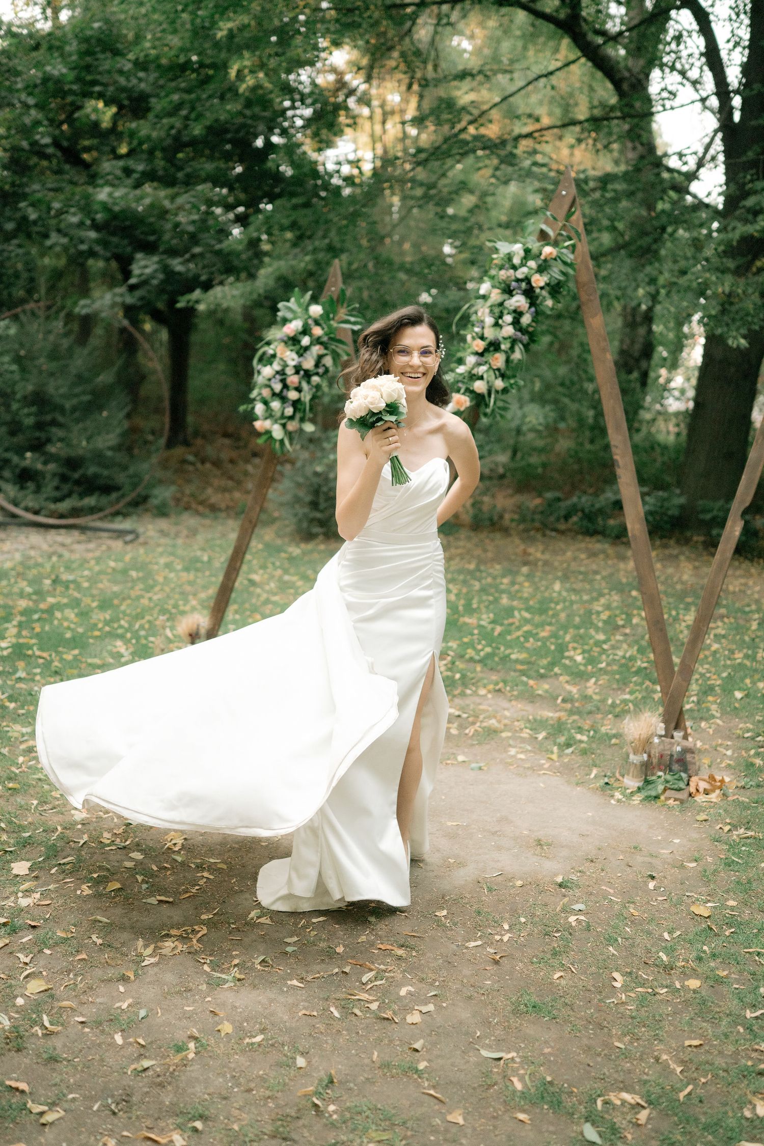 A bride in a flowing white wedding dress twirls playfully in a wooded outdoor setting during a photo session.