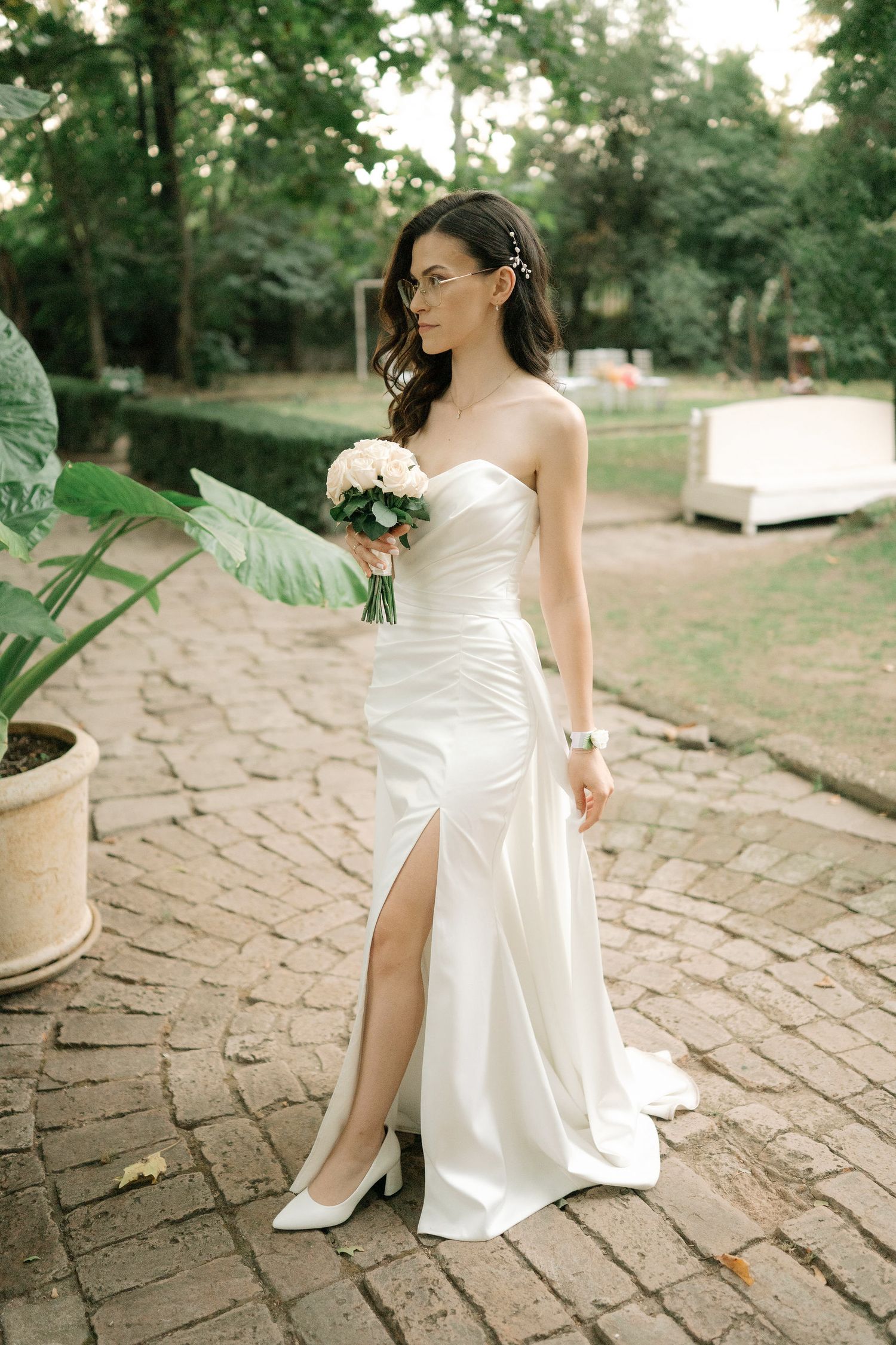 A white satin wedding dress is elegantly displayed on stone pavers in a garden setting.