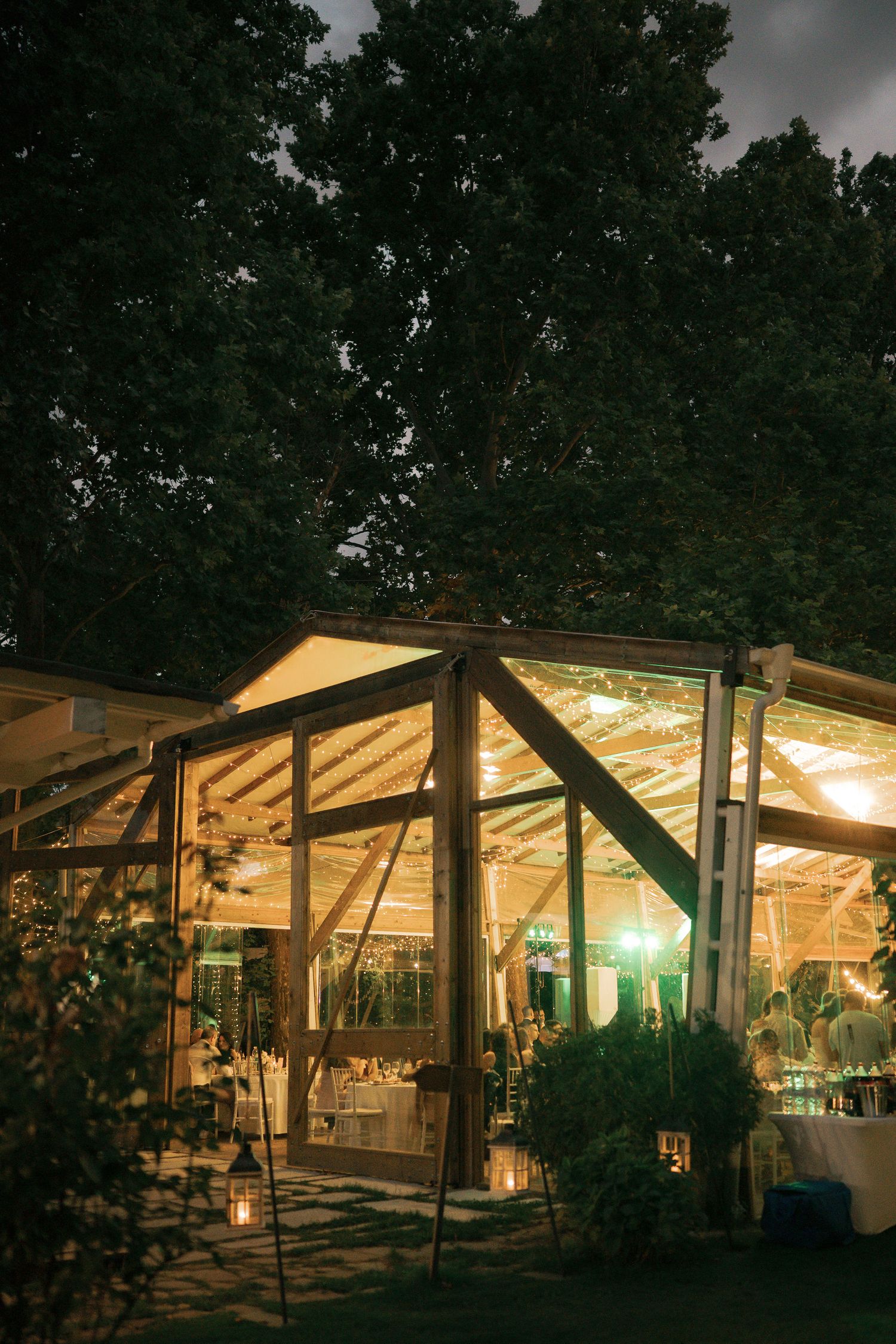A warmly lit outdoor wedding tent glows at dusk with string lights illuminating tables and greenery decorations.