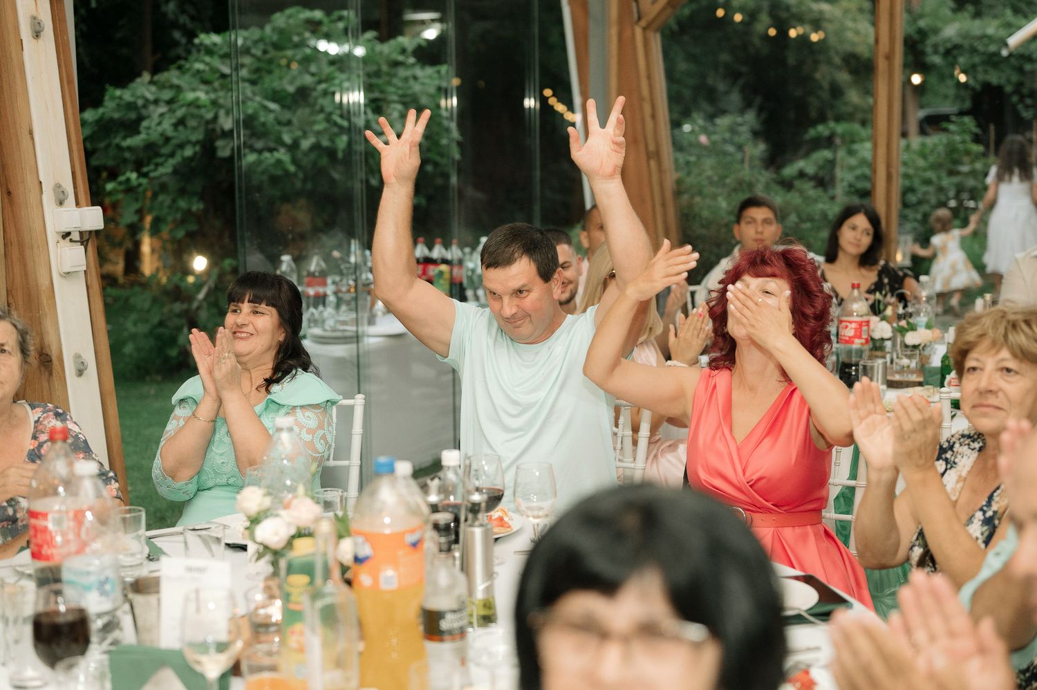 Wedding reception guests raise their hands in celebration at a long dining table with wine glasses.
