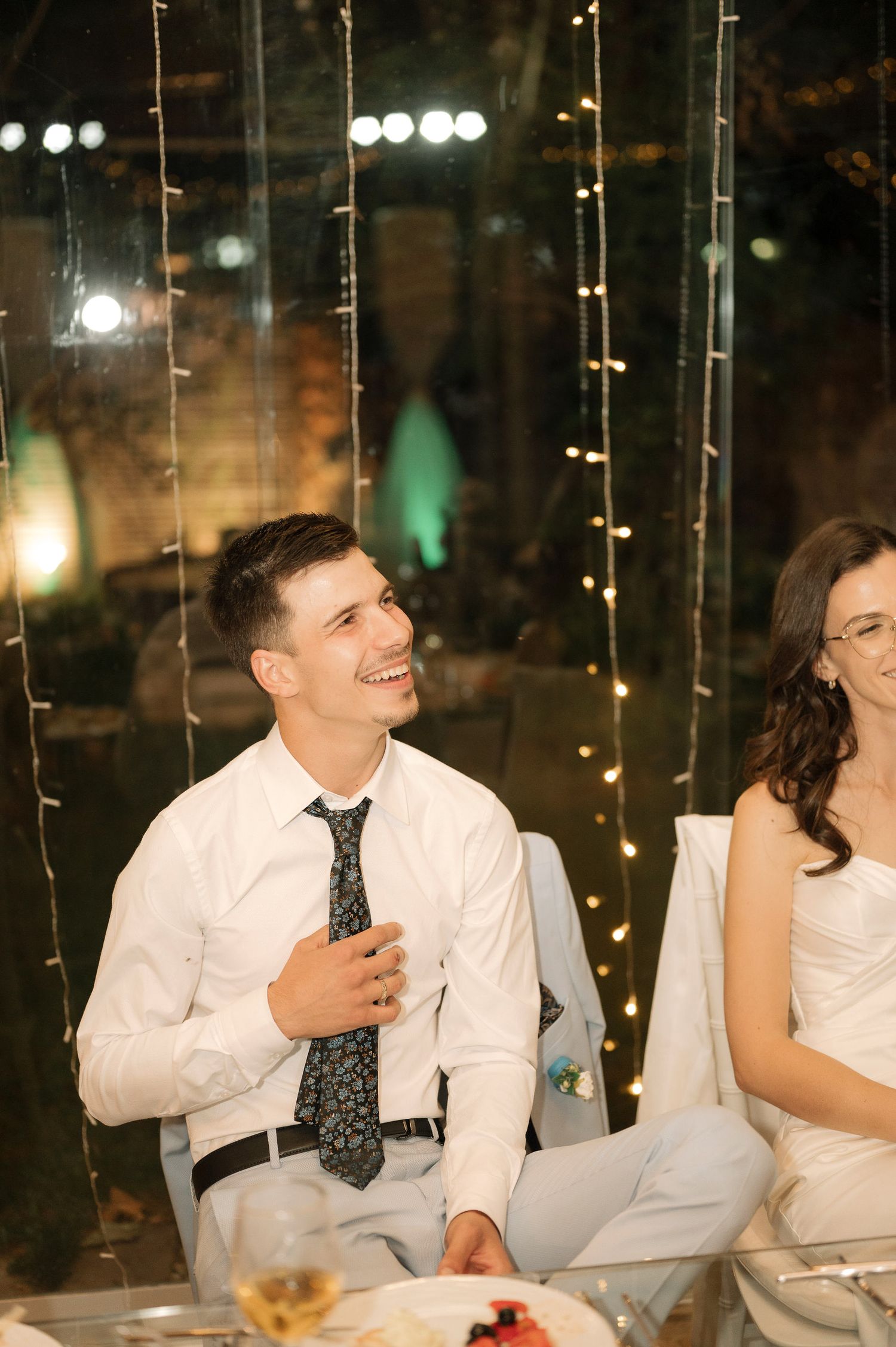 Wedding guests share laughter and smiles while seated at an outdoor reception with twinkling lights in the background.
