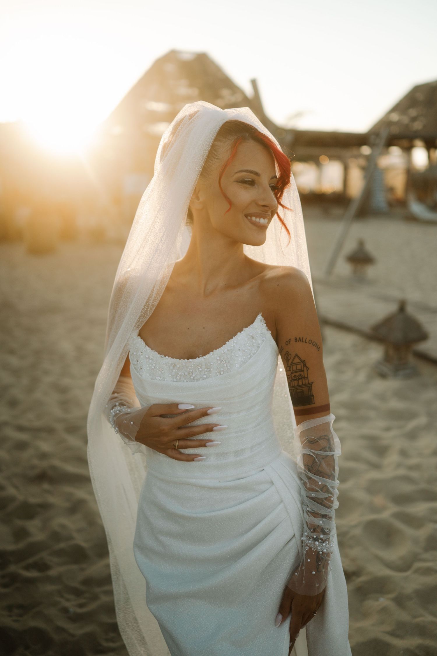 A bride in a white wedding dress and veil poses on a sandy beach at sunset with visible tattoos on her arms.