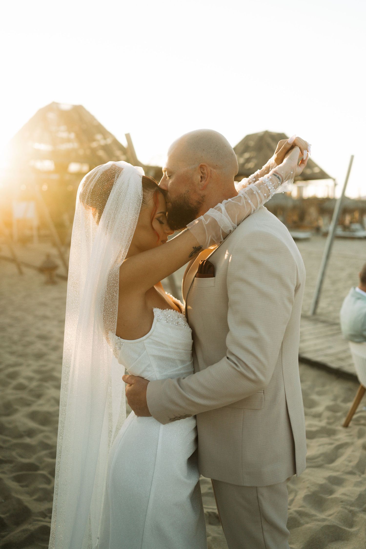 Newlywed couple shares intimate kiss on beach at sunset with bride in white dress and groom in tan suit.
