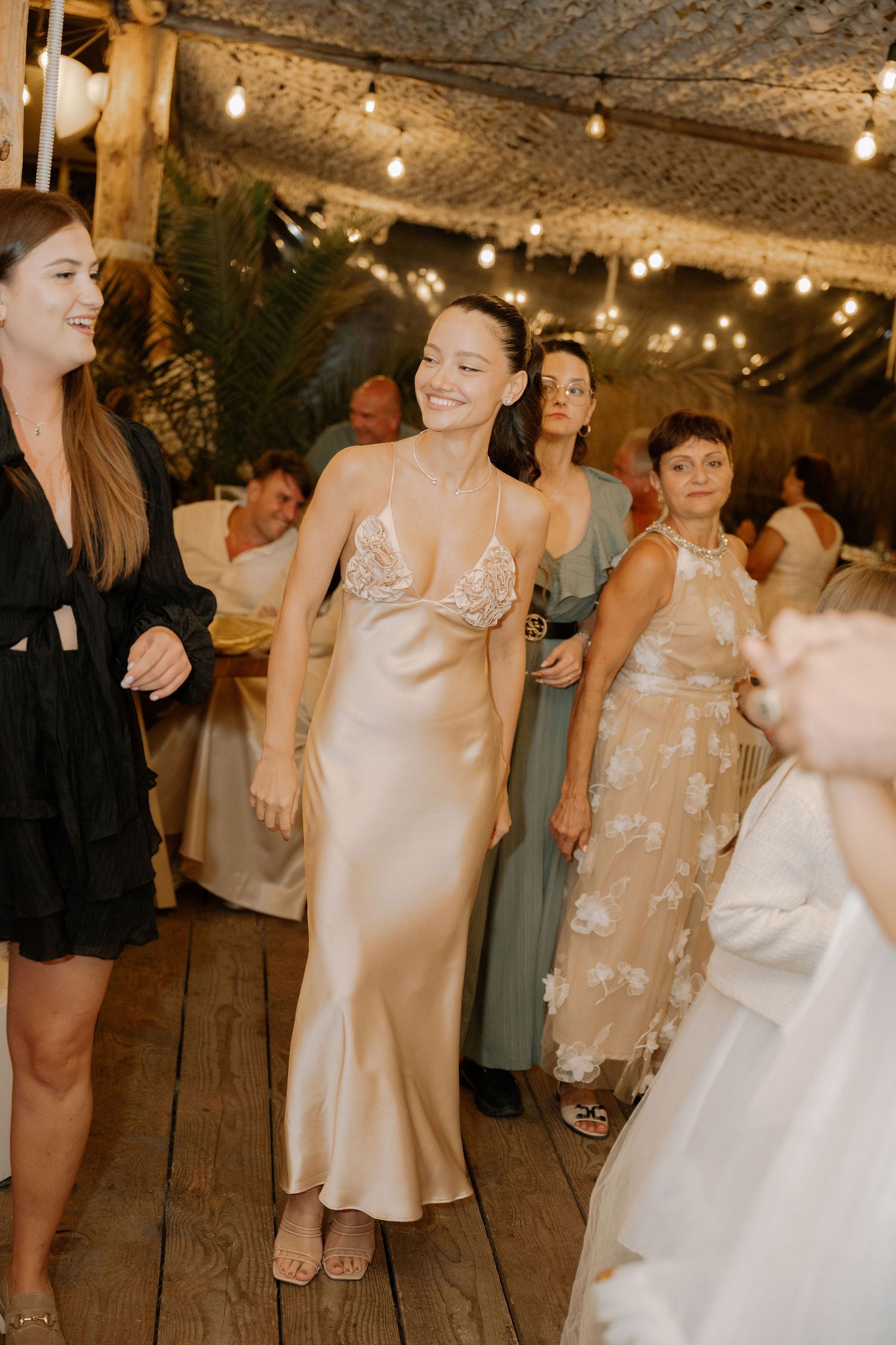 Wedding guests dance and celebrate together on a rustic wooden floor under warm string lights at an evening reception.