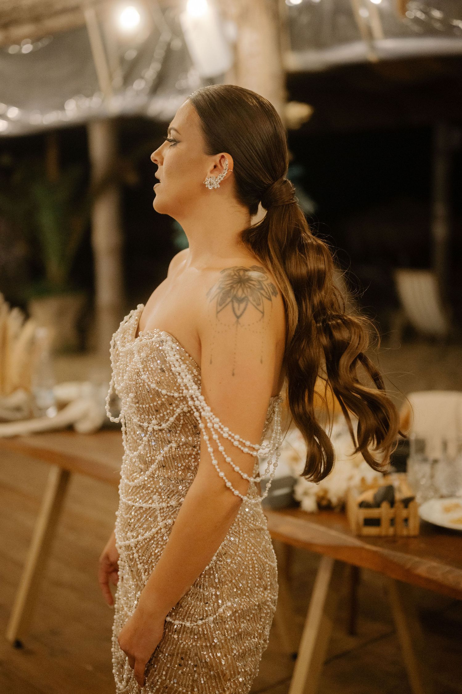A bride in a sparkling beaded wedding dress poses elegantly in a dimly lit reception venue.