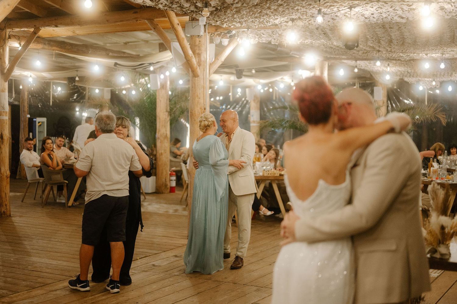 Couples dance together under warm string lights at a rustic outdoor reception venue with wooden beams overhead.