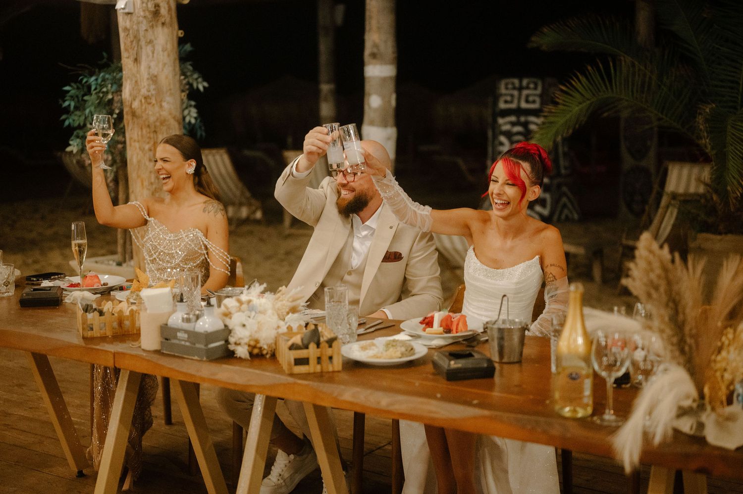 Wedding guests in white attire gather around a rustic wooden table at an outdoor evening reception.