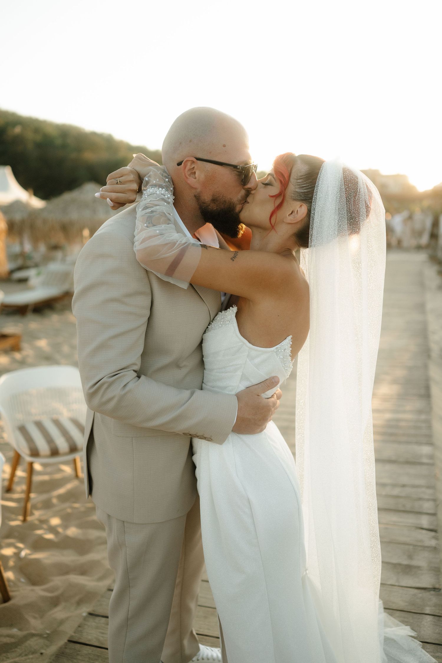 Newlywed couple shares romantic kisses on a sunlit wooden boardwalk at sunset during their beach wedding.