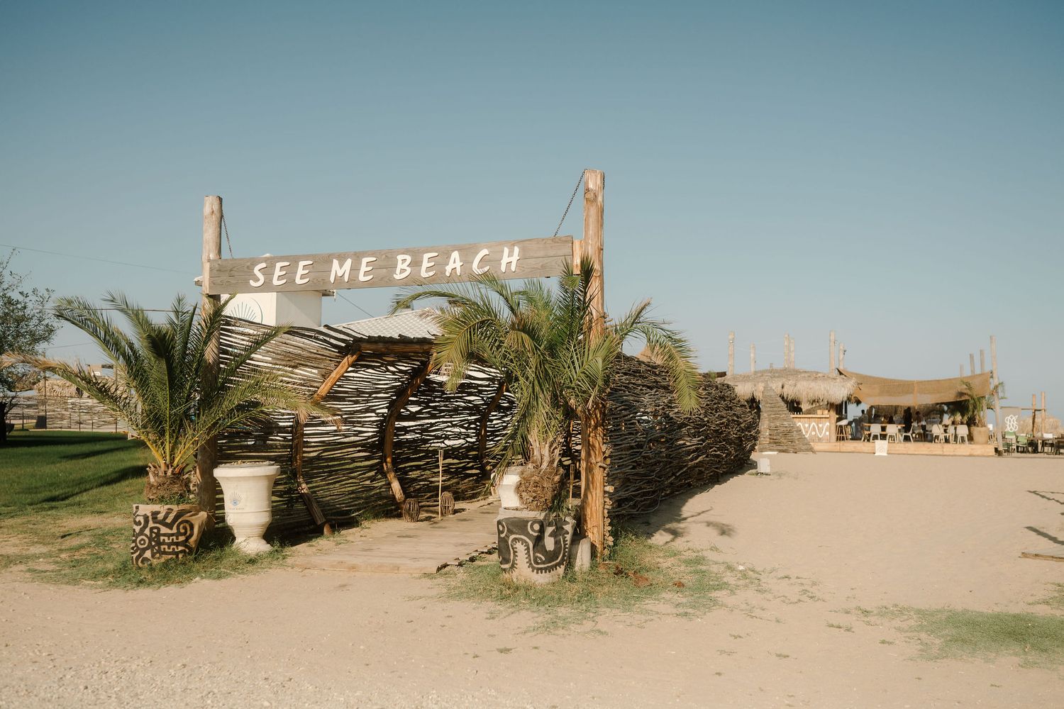 A panoramic view of a rustic wooden beach shack with palm trees and 'See Me Beach' signage in a desert landscape.