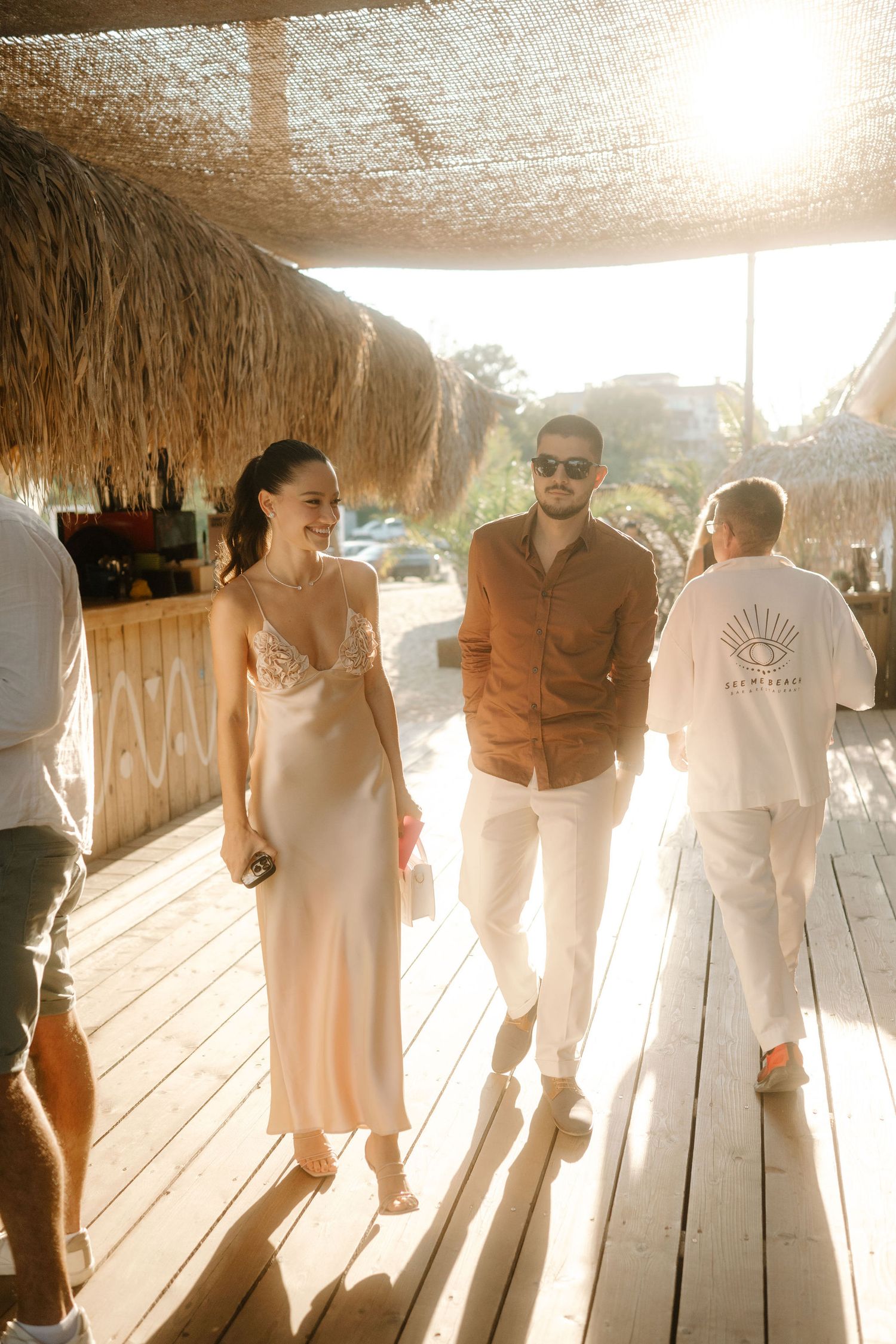 People walking along a wooden deck under a thatched roof with bright sunlight creating silhouettes and shadows.