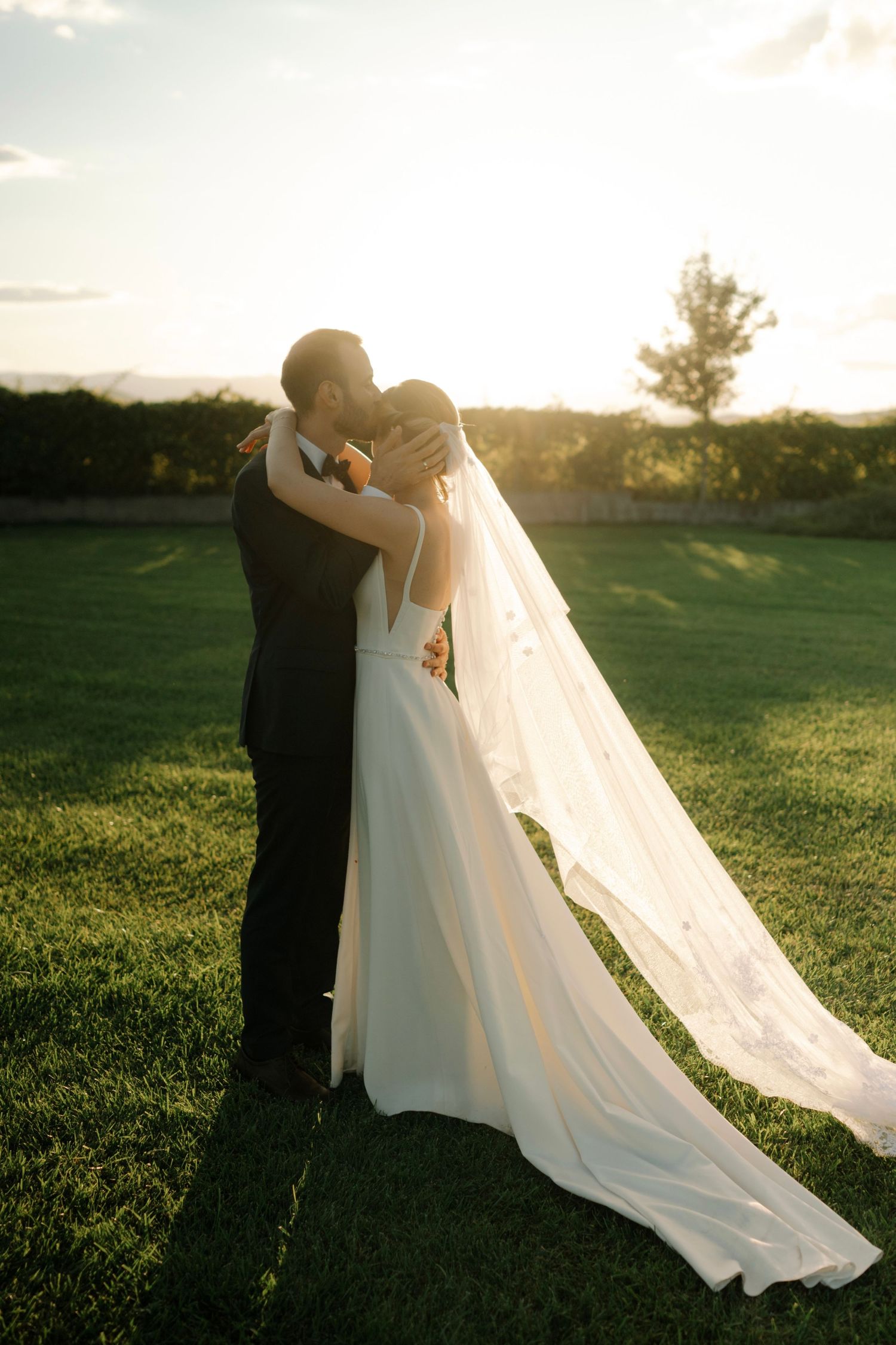 A couple embraces during golden hour in a grassy field with the bride's flowing white veil trailing behind them.
