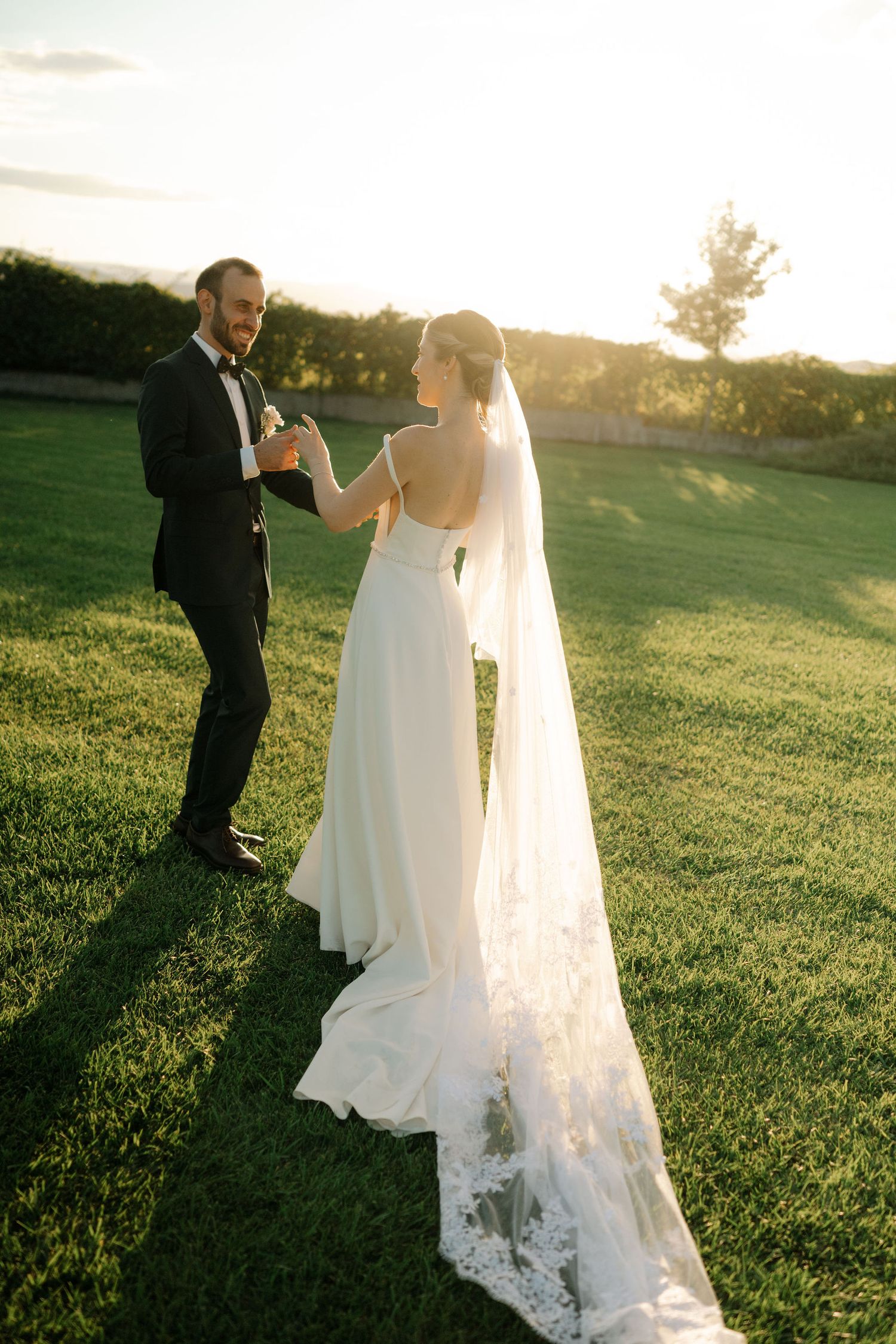 A couple in wedding attire walks together on a grassy hillside during golden hour sunset.
