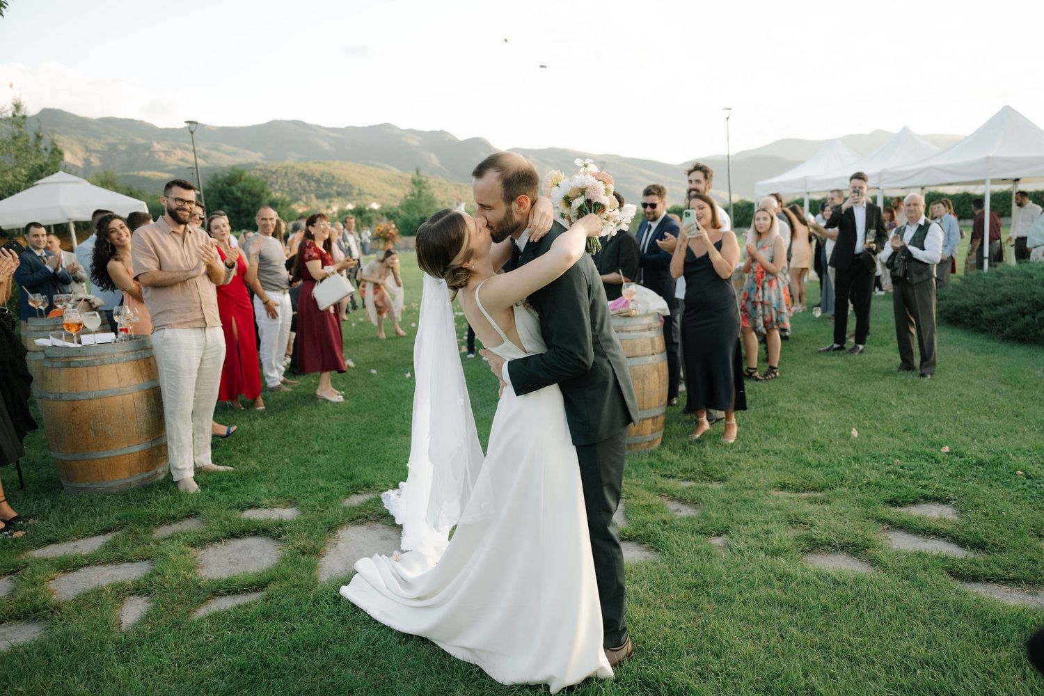 Guests gather on a scenic hillside vineyard for an elegant outdoor wedding celebration with wine barrels and mountain views.