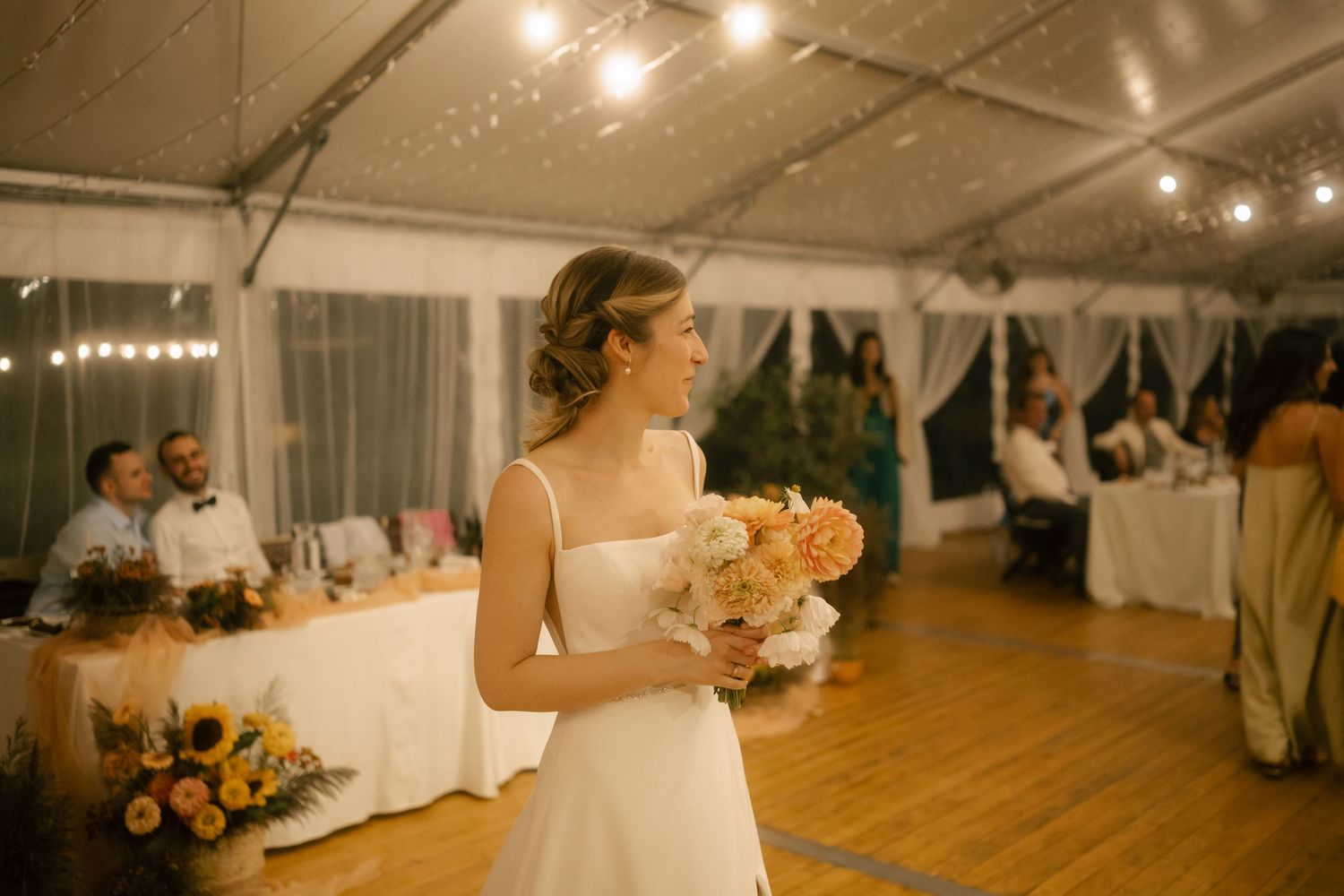A romantic wedding reception under a white tent with glowing lights illuminating the dance floor and dining tables.