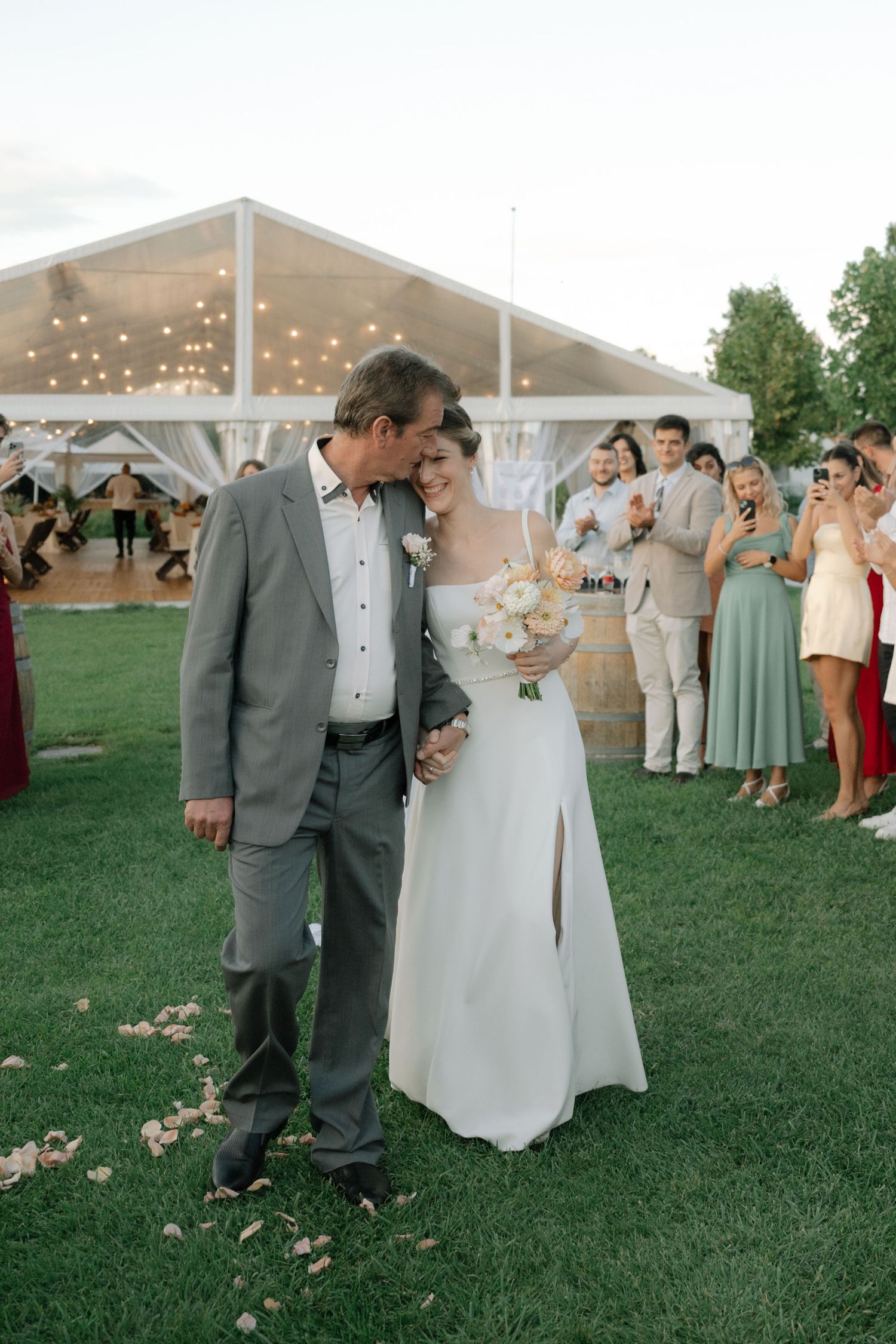 Wedding guests mingle and celebrate under an elegant outdoor tent on a lush green lawn at sunset.