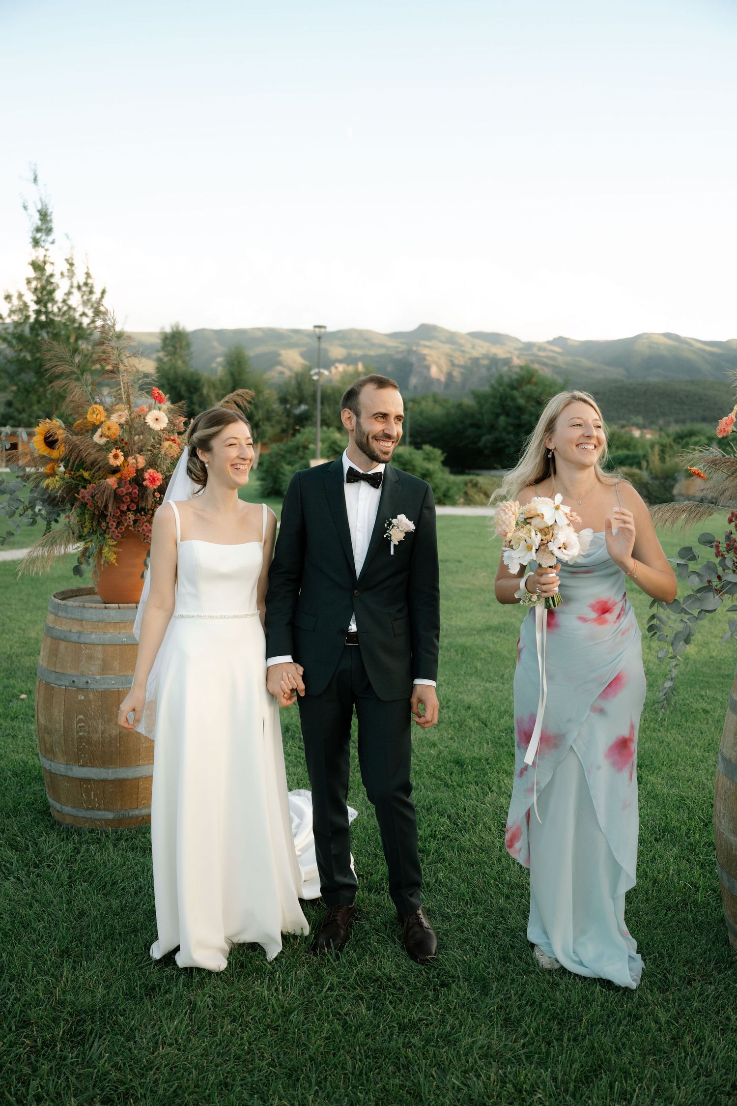 Wedding party members pose together on a grassy hillside with wine barrels and floral decorations at sunset.