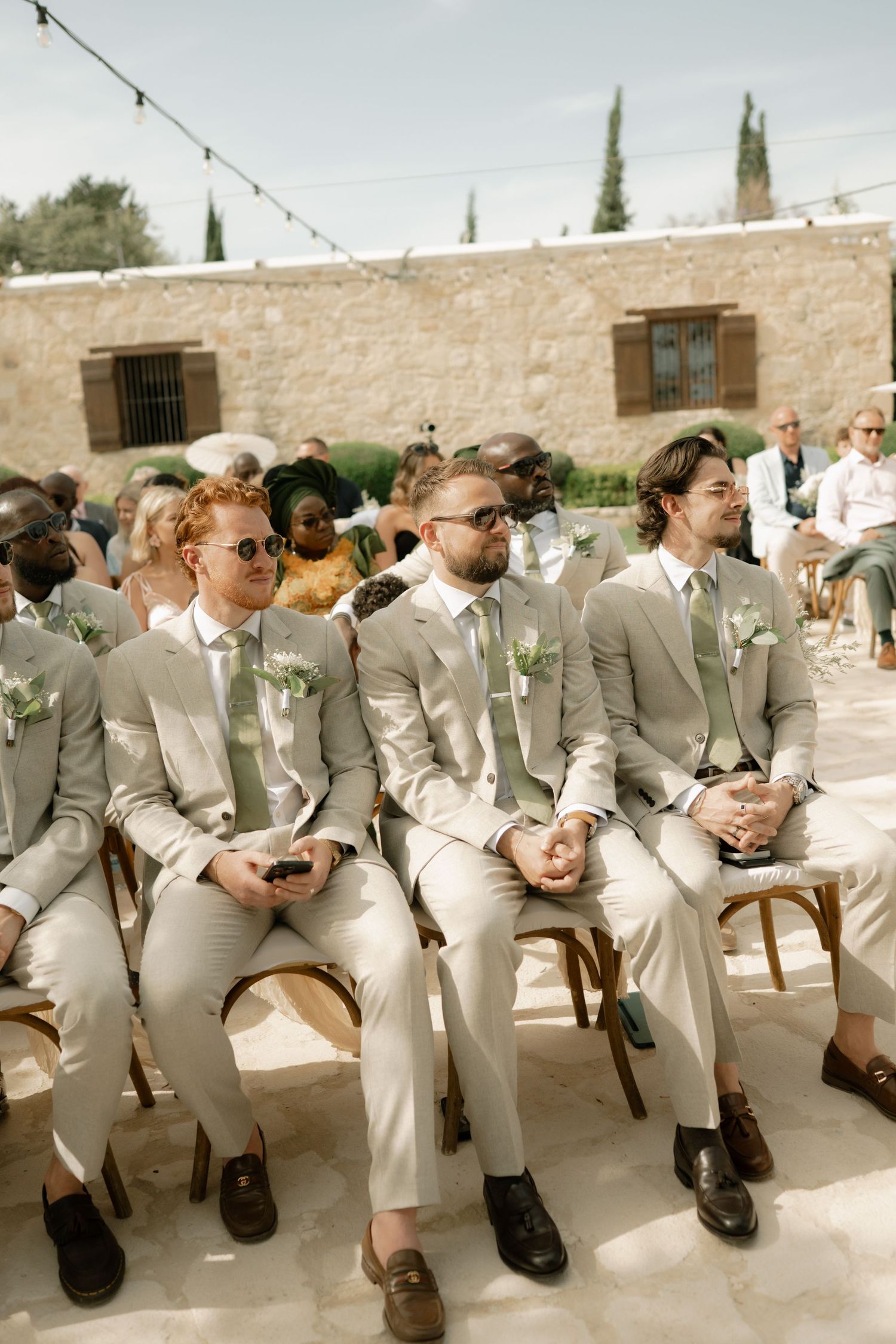 Groomsmen in matching sage green suits with floral boutonnieres seated at an outdoor wedding ceremony.
