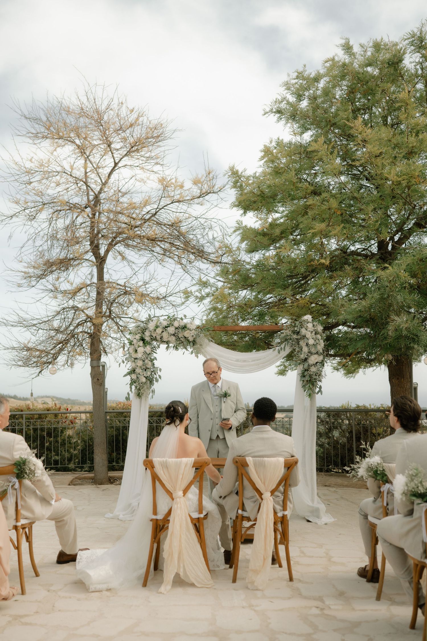 Outdoor wedding ceremony with couple at floral arch, officiant, and guests seated on wooden chairs amid trees.