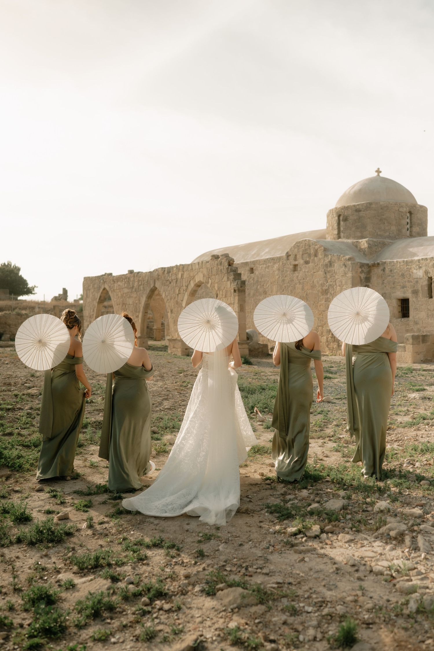 Bride and bridesmaids in sage green dresses holding white parasols near ancient stone ruins.