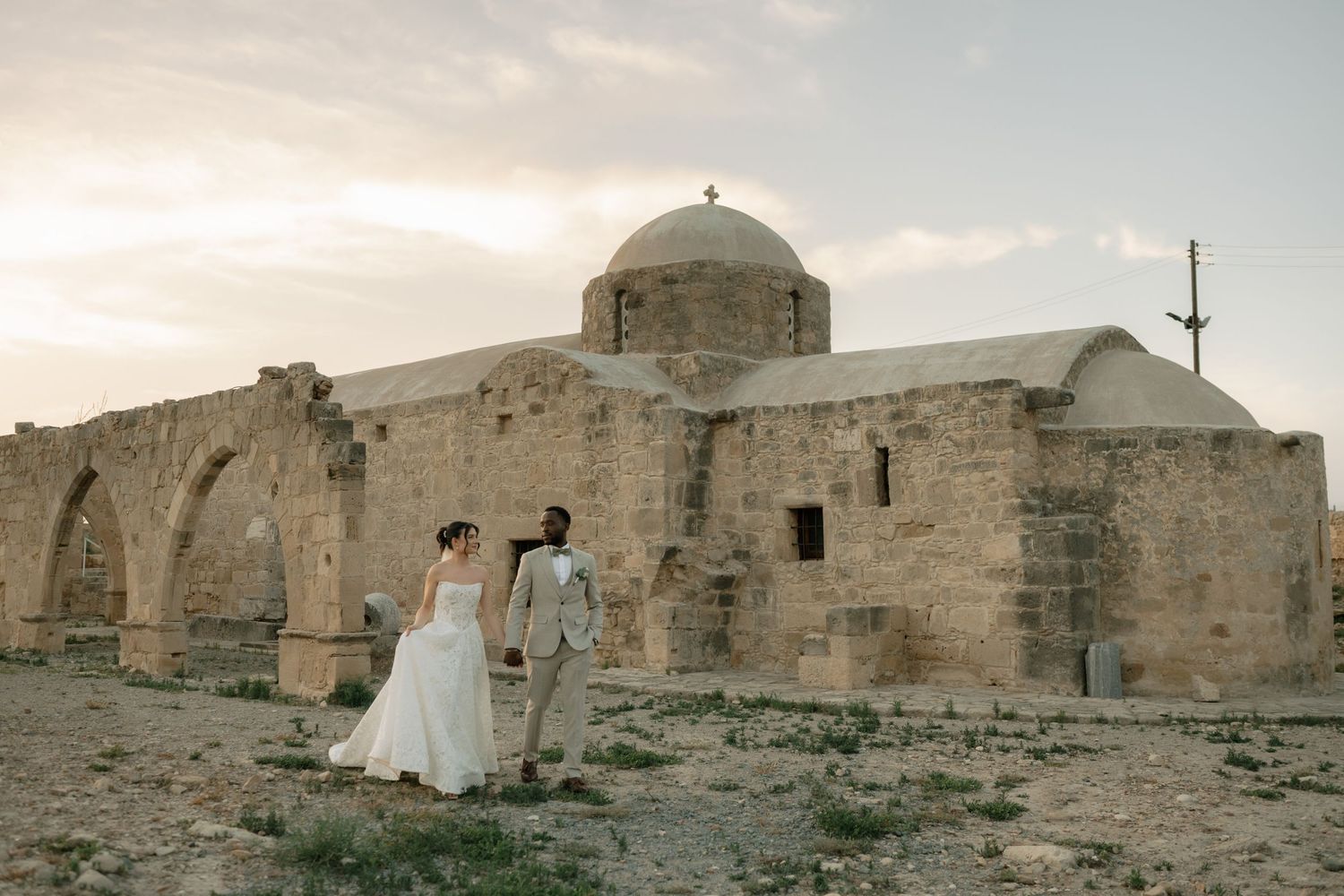Bride and groom walk hand-in-hand before a historic stone church at sunset in Cyprus.