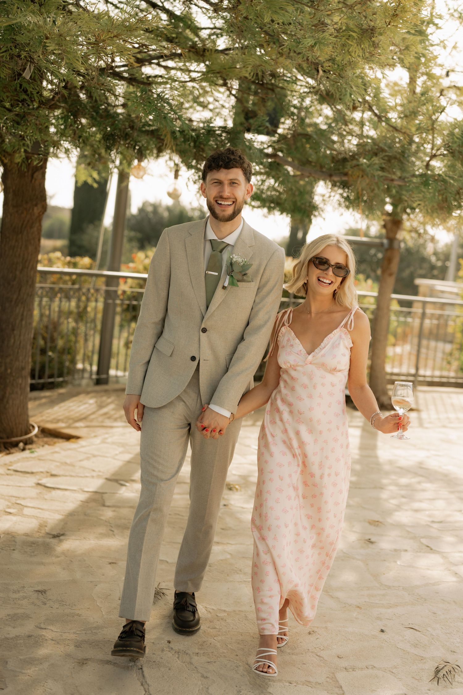 A smiling couple walks hand in hand outdoors, man in tan suit, woman in floral dress and sunglasses.