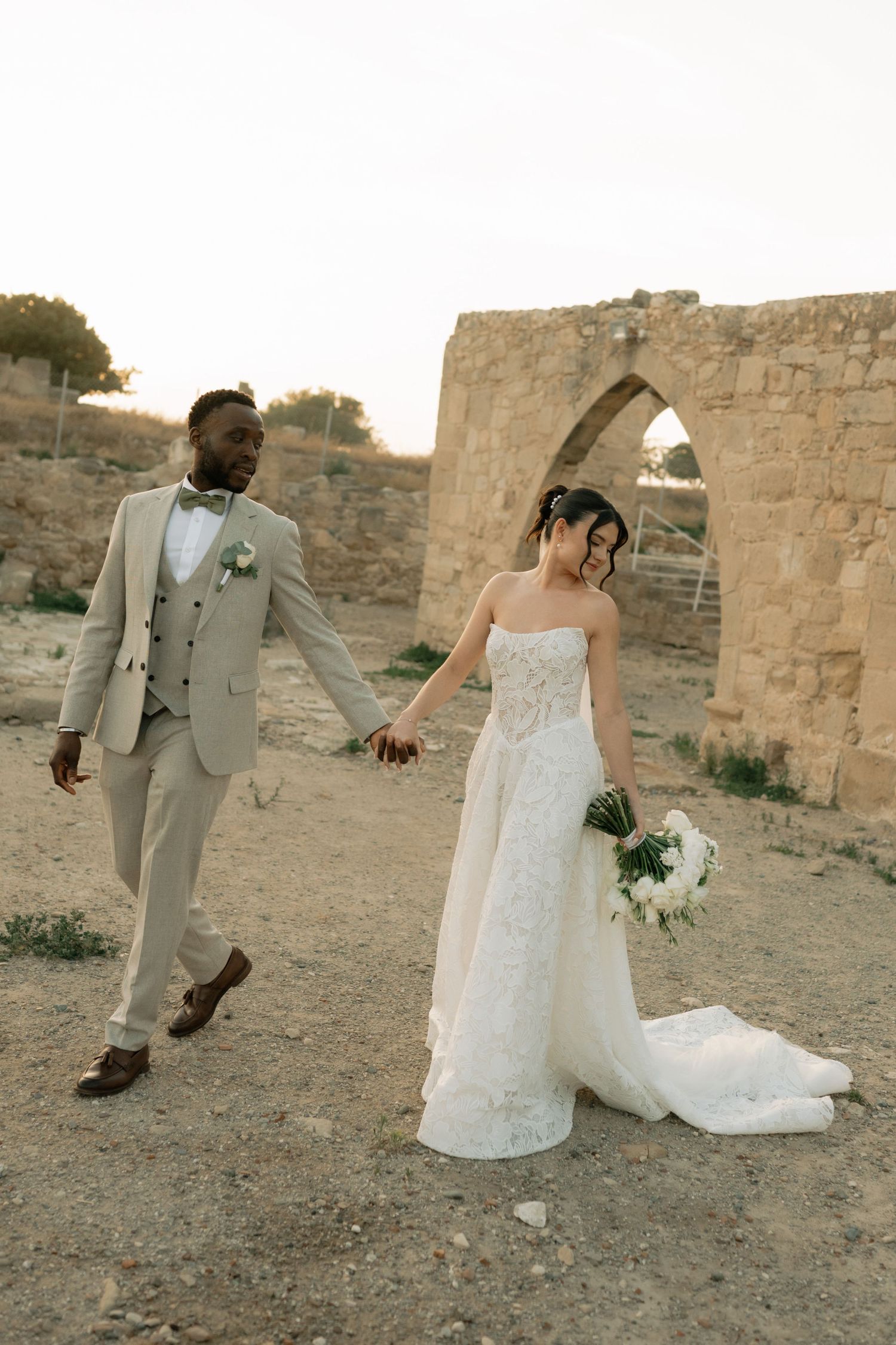 Interracial couple in wedding attire walking hand in hand near ancient stone ruins at sunset.
