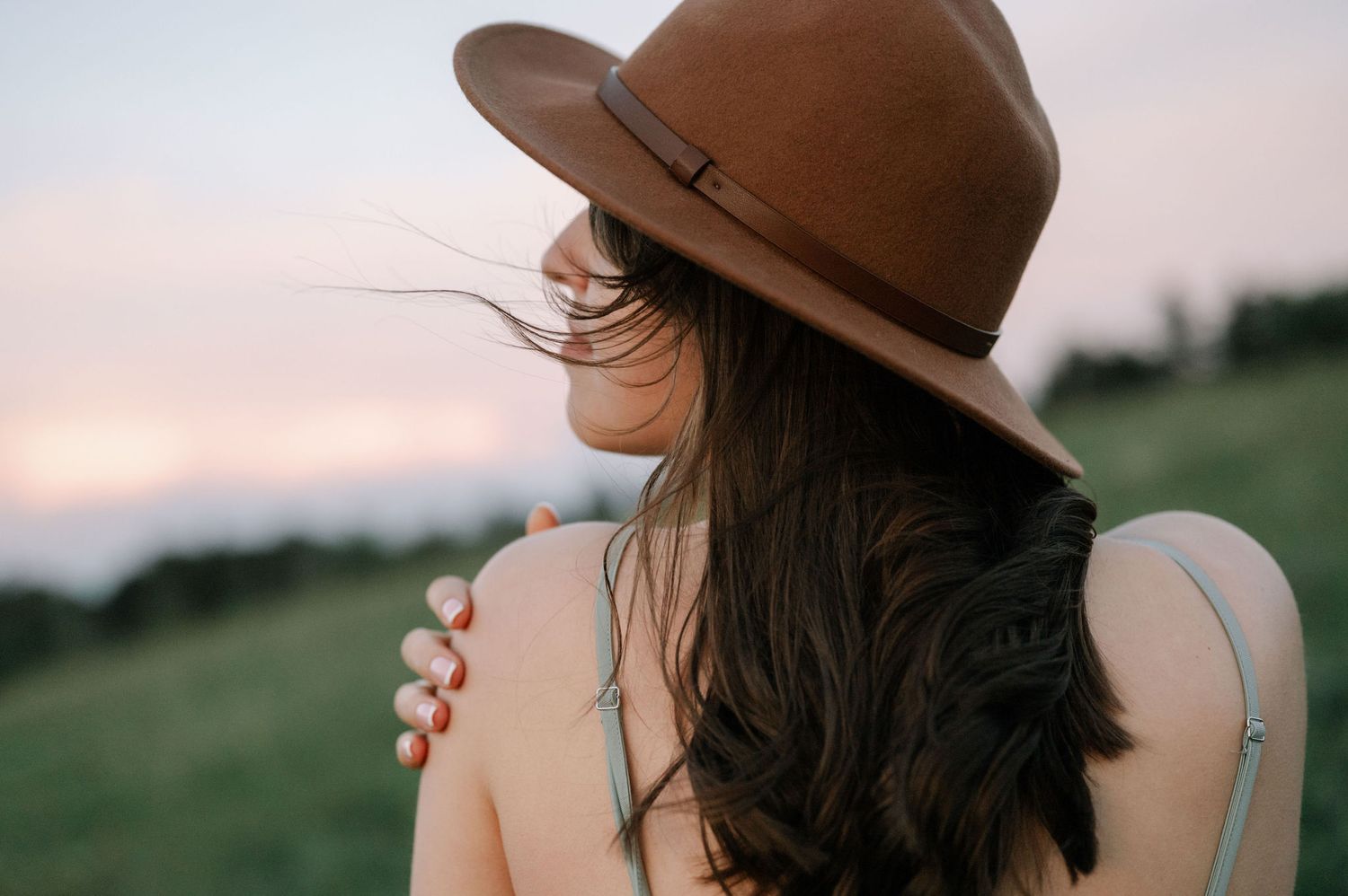 Woman in brown felt hat with dark hair, viewed from behind, against a scenic outdoor sunset backdrop.