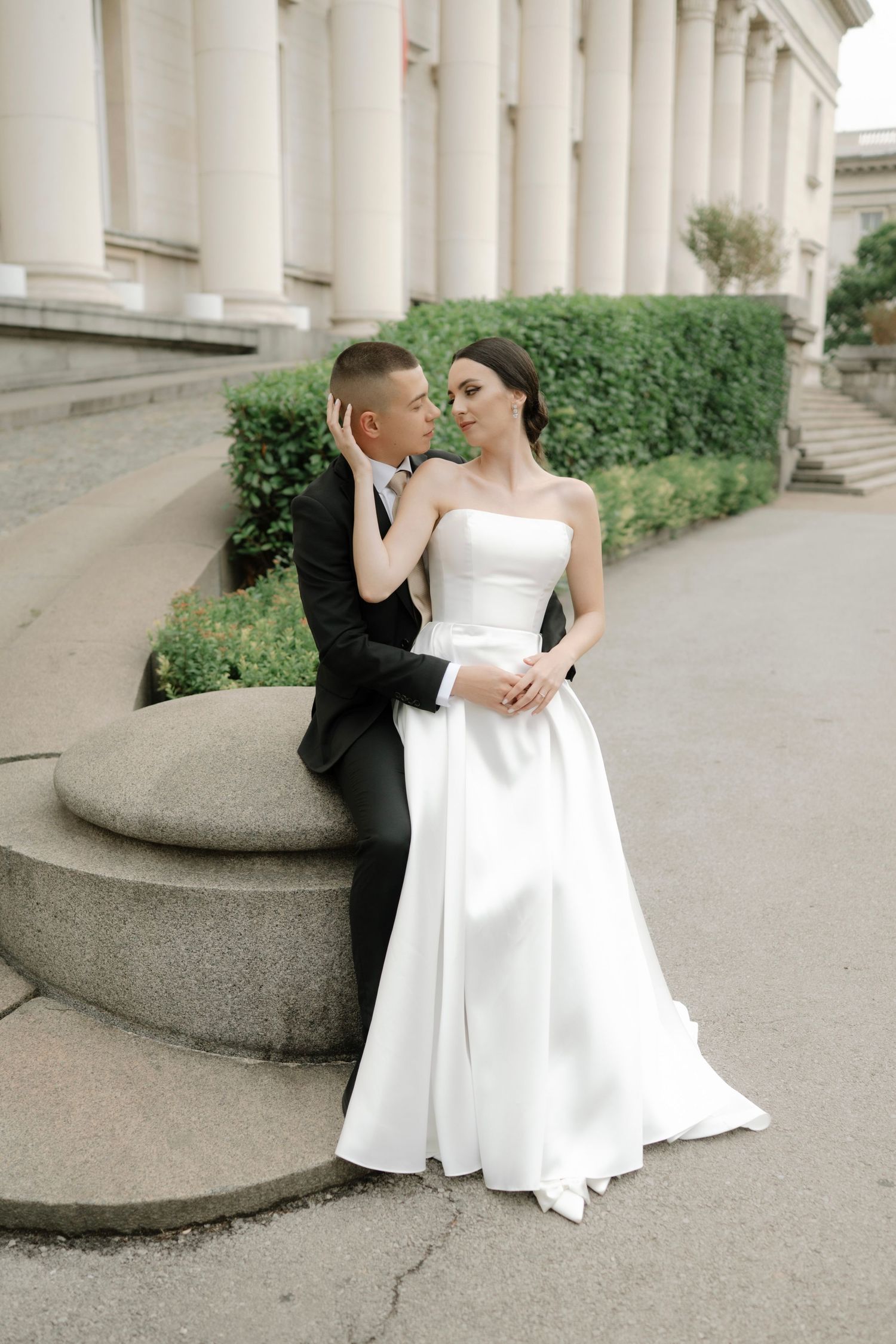 A couple in formal wedding attire poses together on stone steps in front of a classical building with white columns.