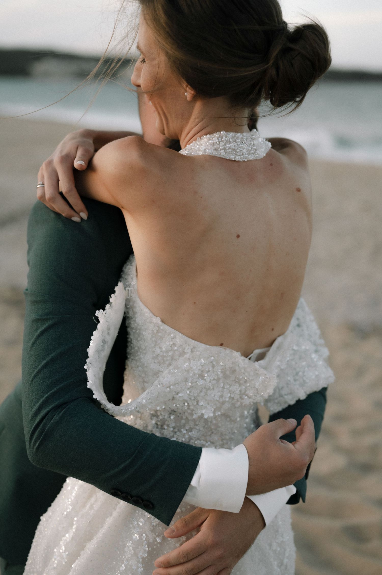 Bride in backless beaded gown embraces groom on beach at sunset.
