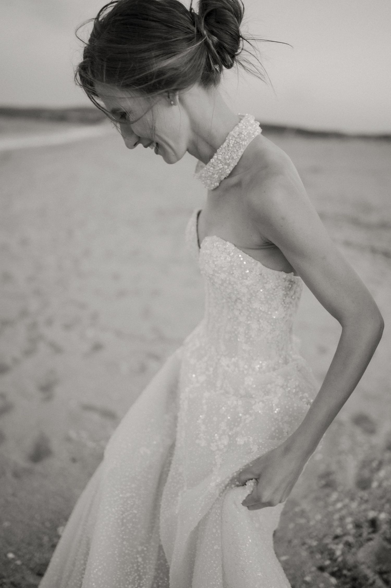 Black and white photo of a bride in a sequined strapless gown with halter neckline, posing on a sandy beach.