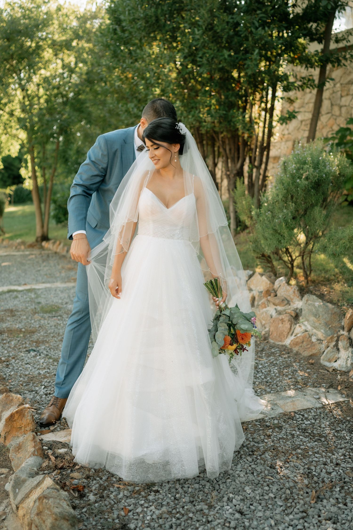 A bride in a flowing white tulle gown and veil holds a bouquet on a gravel path surrounded by greenery.