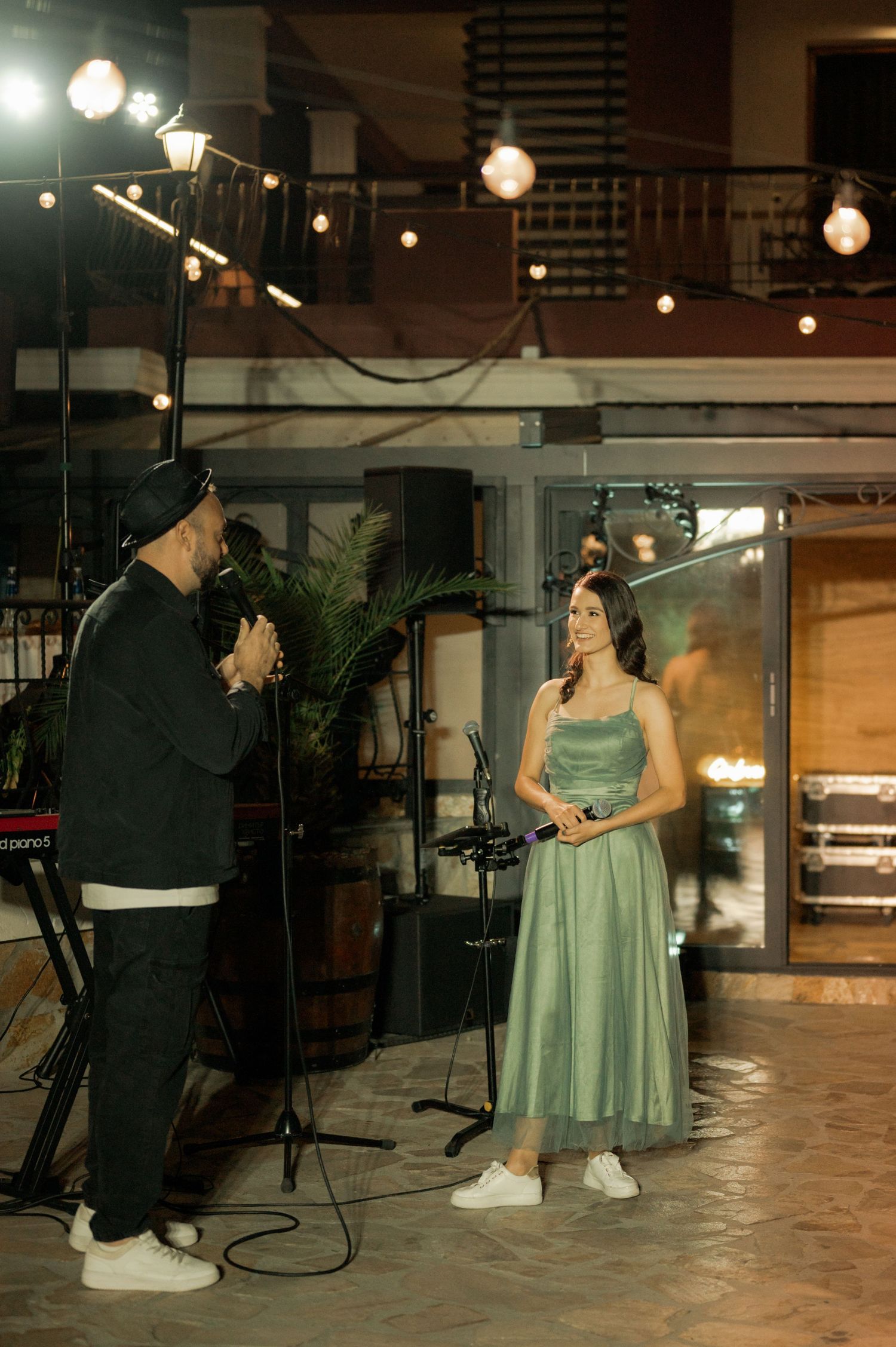 A performer in a black outfit and someone in a mint green formal dress stand under string lights at an indoor venue.