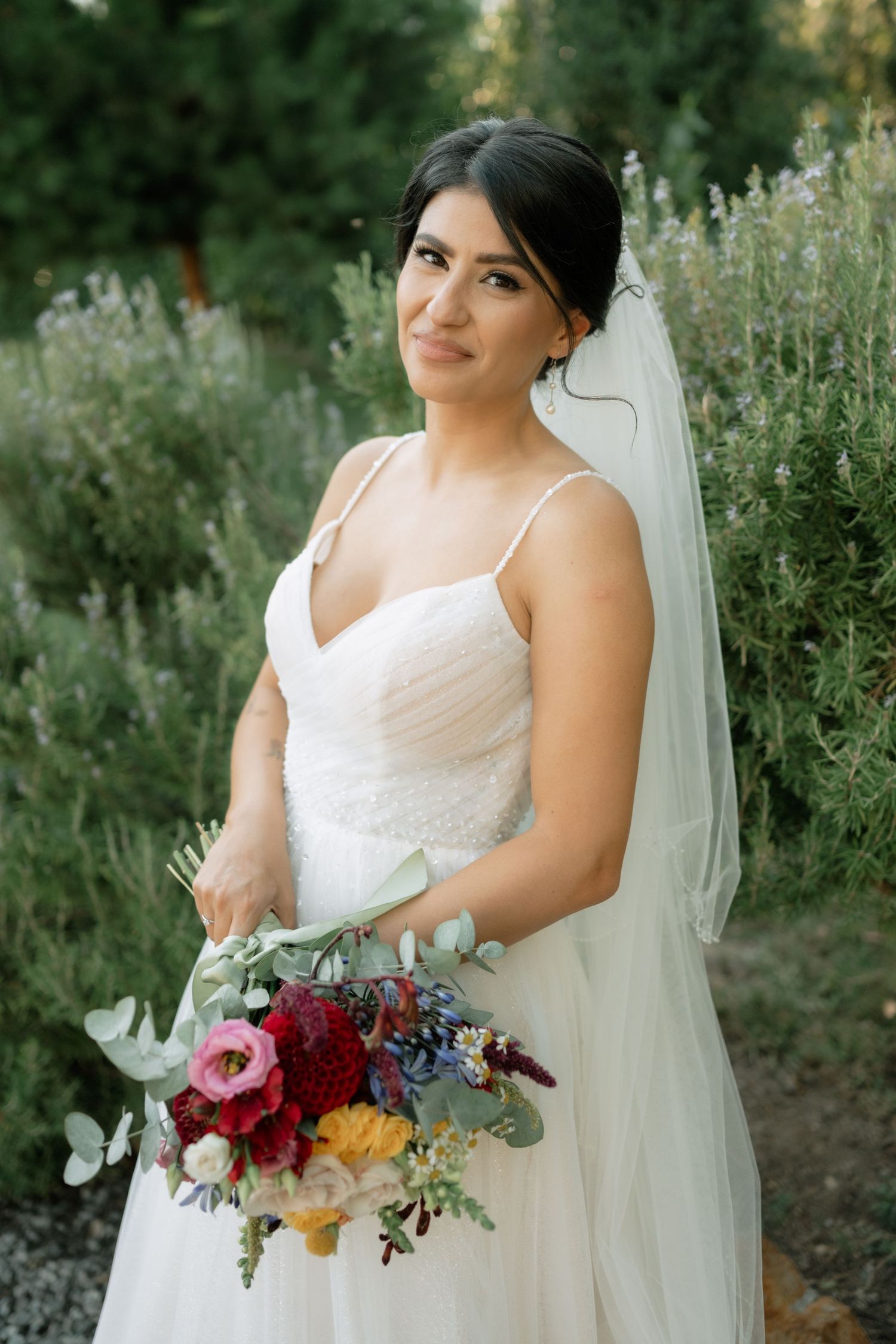 Bride in white wedding dress holds vibrant burgundy and orange bouquet with eucalyptus in outdoor garden setting.
