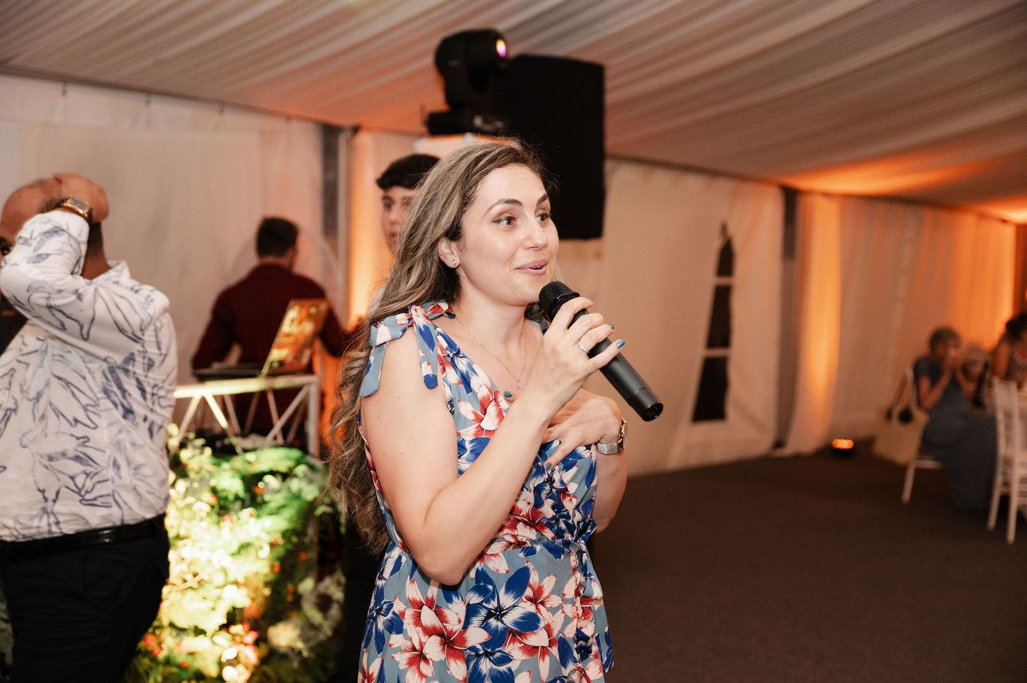 A person in a floral dress speaks into a microphone during an indoor reception event with white draping and decor.