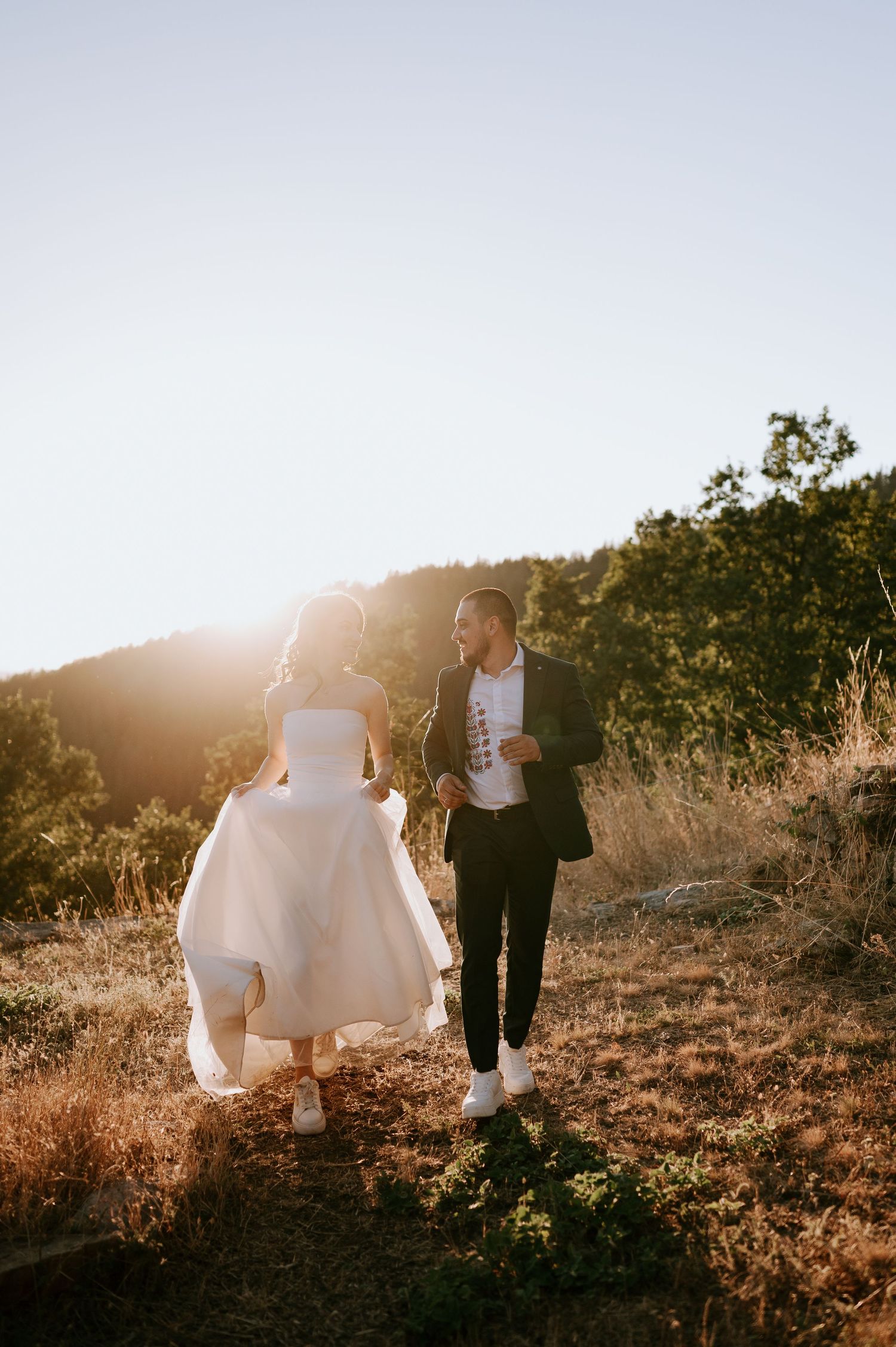 Bride and groom walk hand in hand through golden mountain terrain at sunset.
