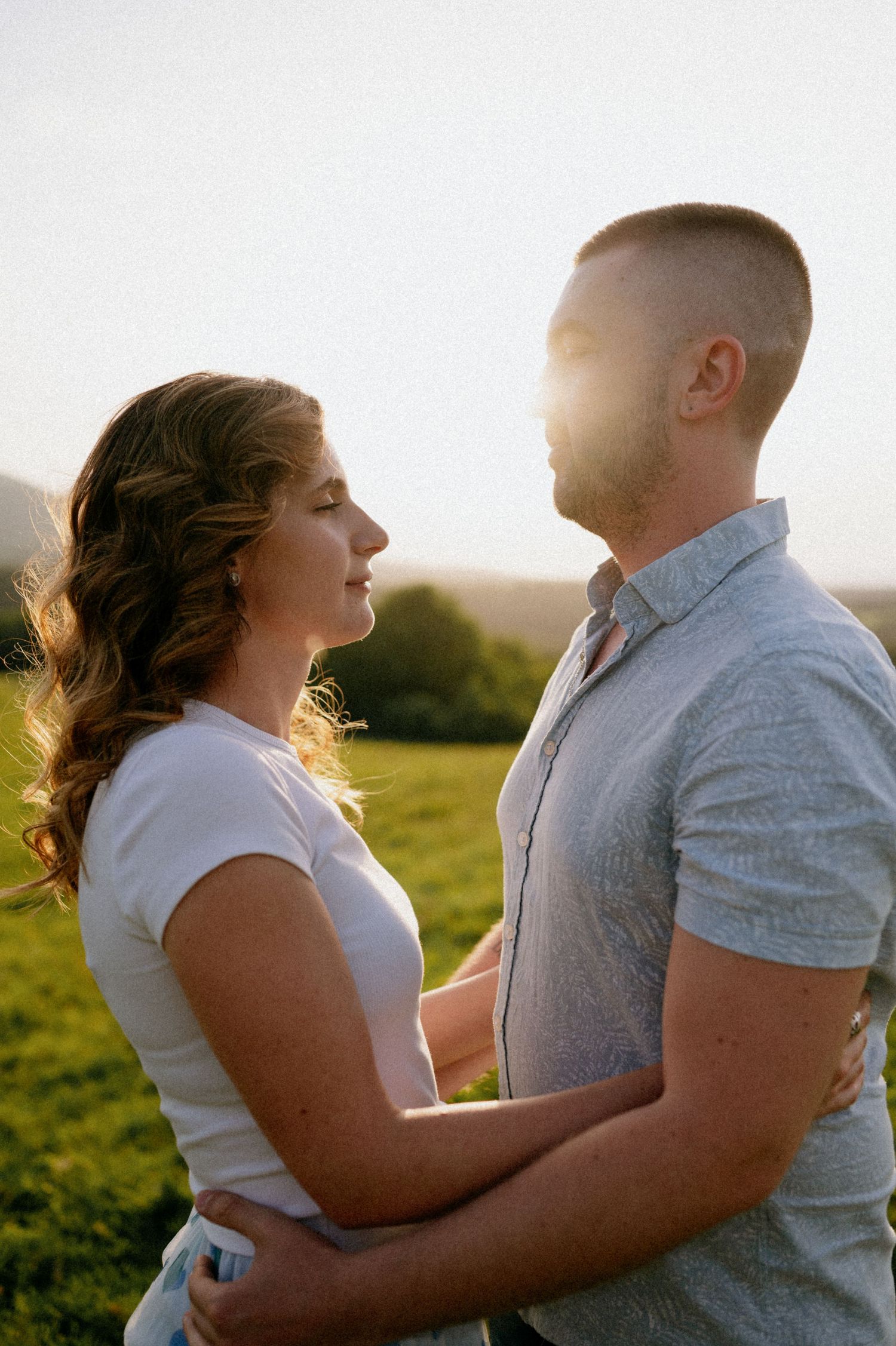 Two people share a tender moment together in a sunlit field with mountains in the background.