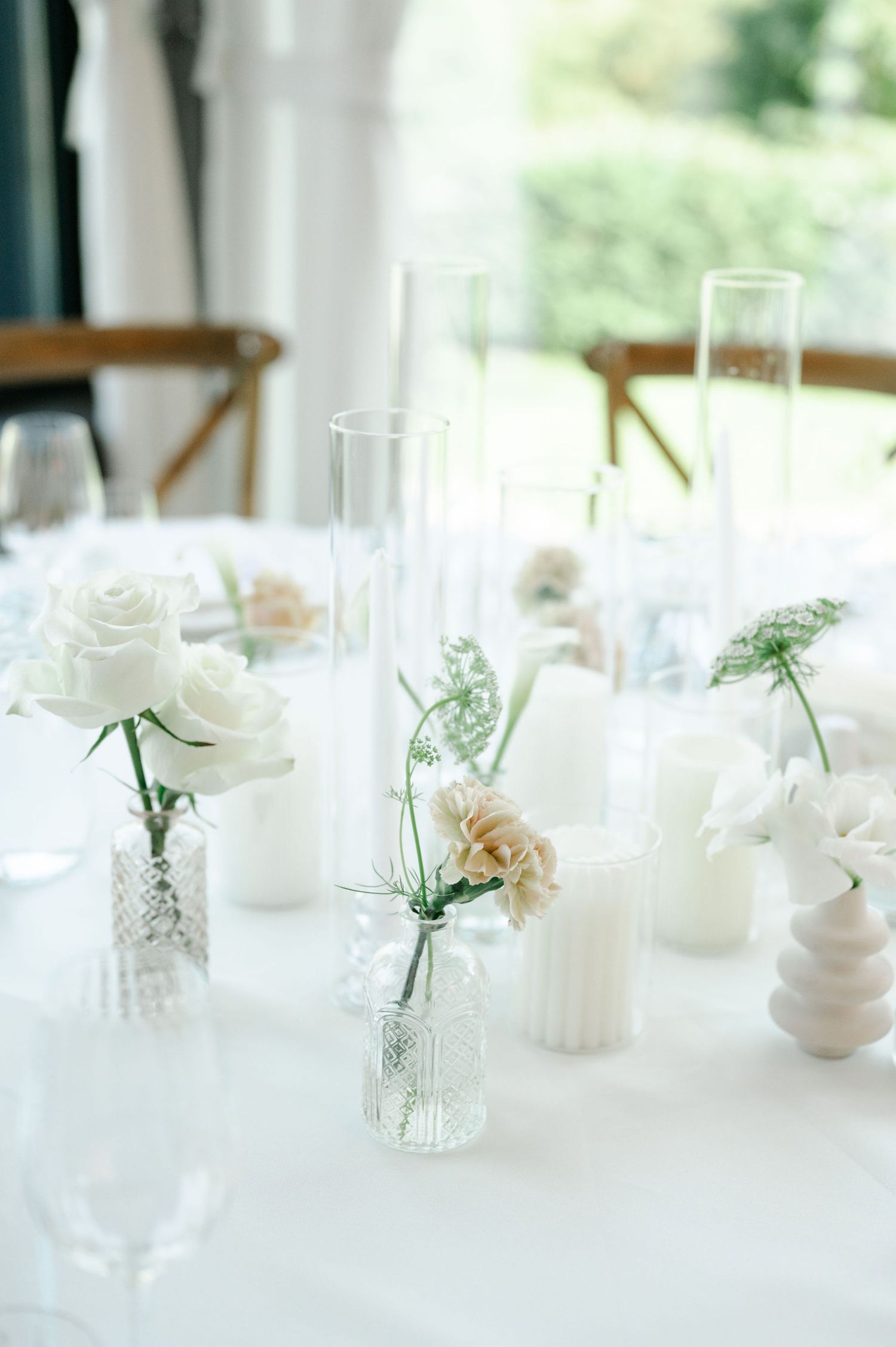 Small white floral centerpieces in glass vases arranged along dining tables with natural light.