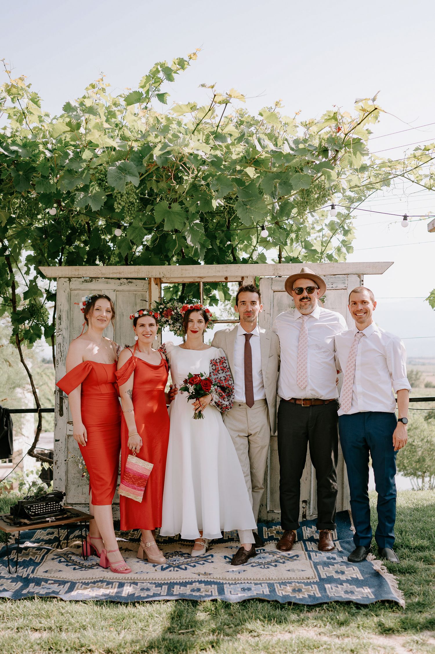 Wedding group photo with bridesmaids in coral dresses and groomsmen under a vine-covered pergola.