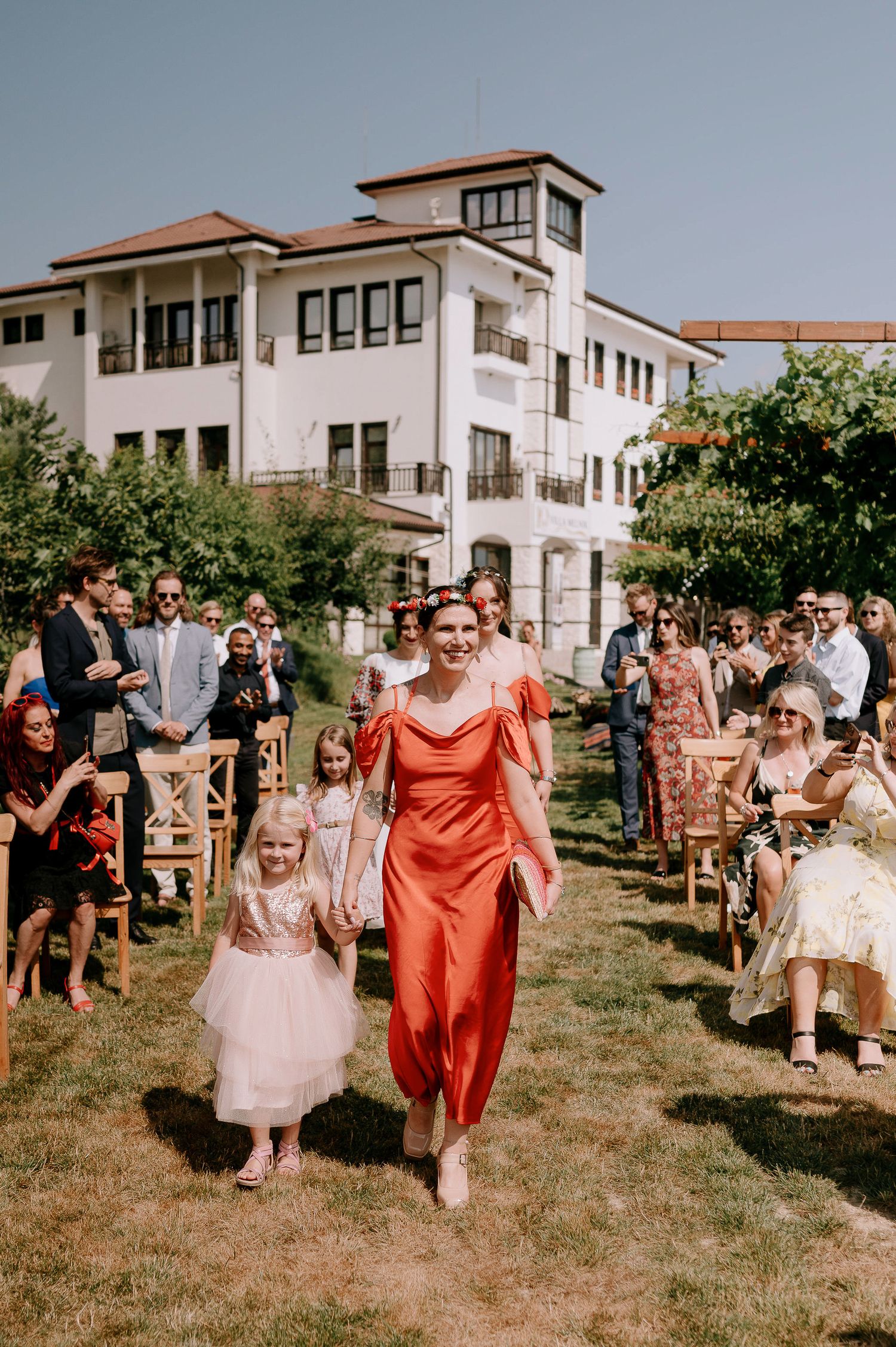 Wedding party members in coral and white dresses walking down a grassy aisle between wooden chairs.