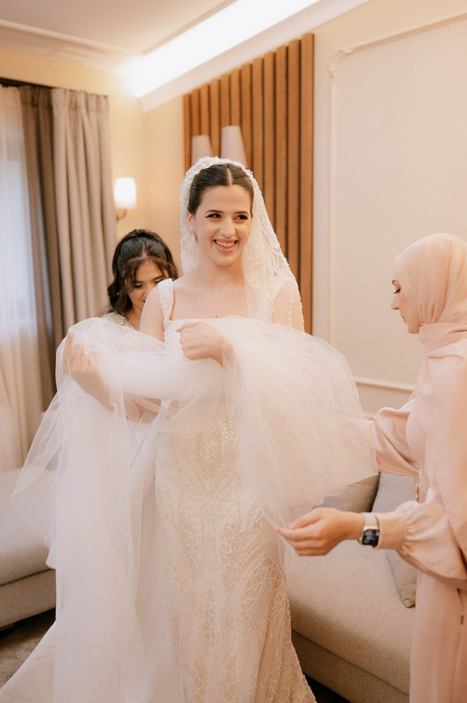 Wedding dress fitting scene with helpers adjusting the bride's gown and veil.