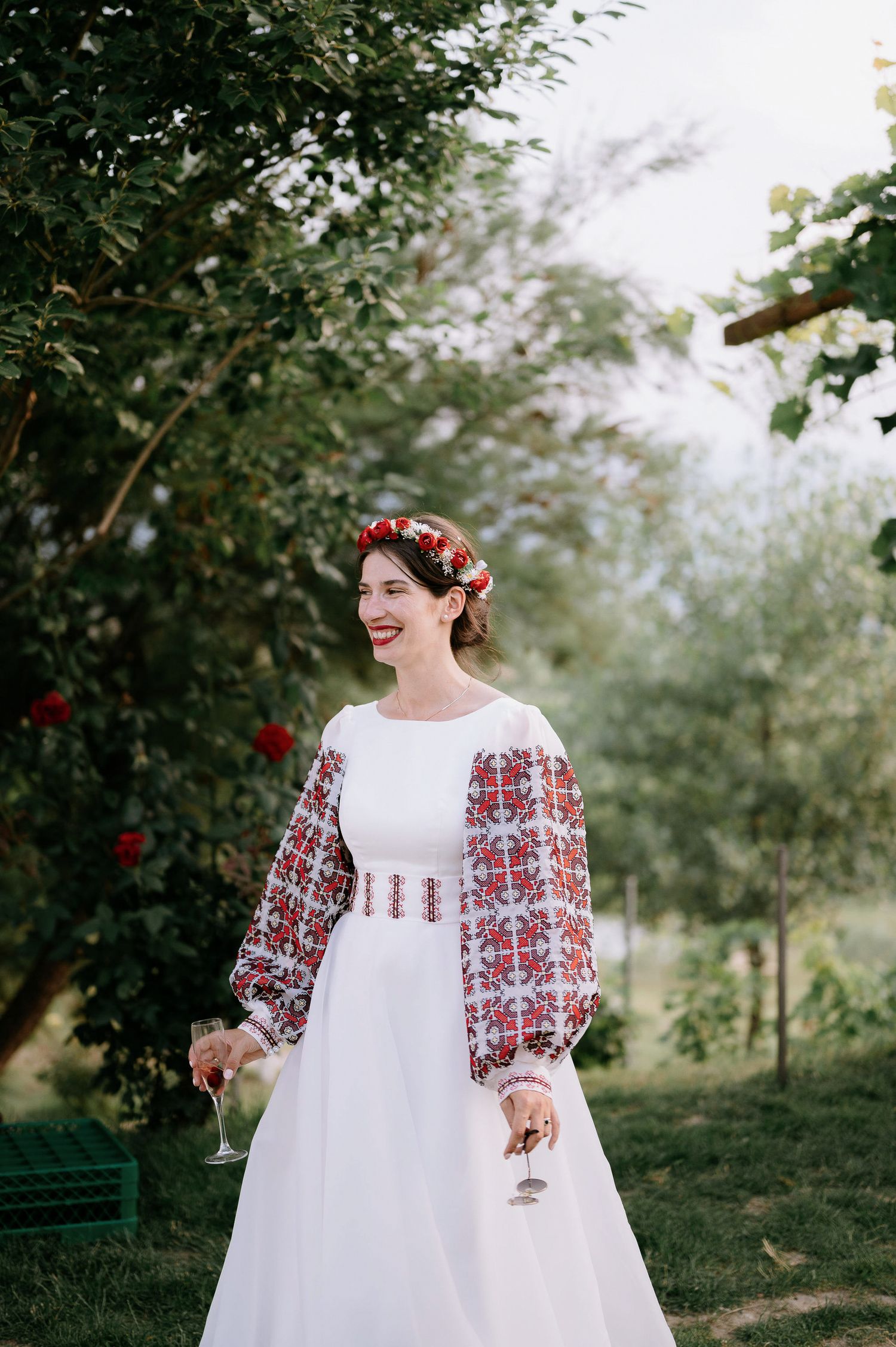 A white dress with red folk-style embroidery and matching headpiece photographed in a garden setting.