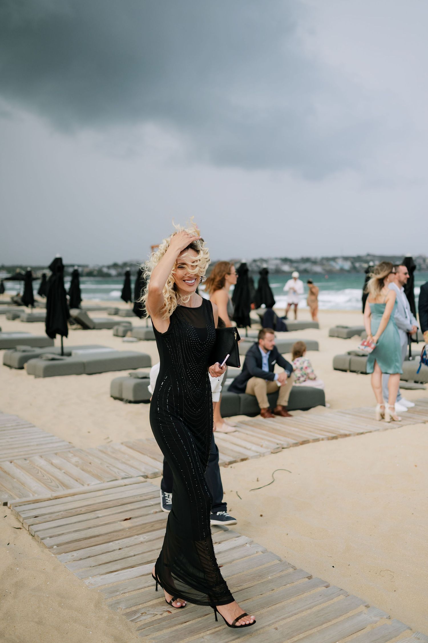 Person in black dress walking along wooden boardwalk at beach wedding.